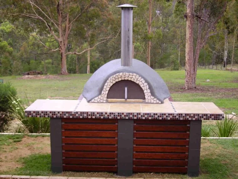 Outdoor pizza oven with stone arch, wooden front, and chimney, set against a backyard with trees and grass.