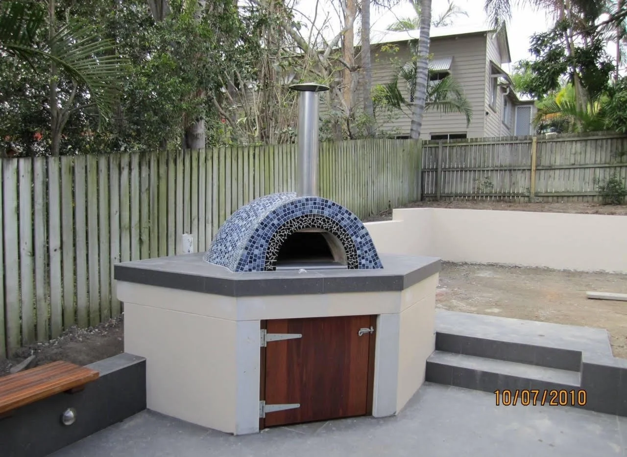 An outdoor brick pizza oven with a mosaic-tiled dome and a chimney, situated on a concrete patio with a wooden door at the front, next to stairs and a wooden fence.