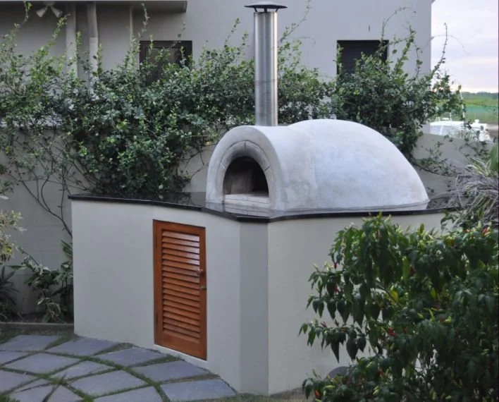 Outdoor pizza oven with a domed top, chimney, and wooden vent door, surrounded by greenery and paving stones.
