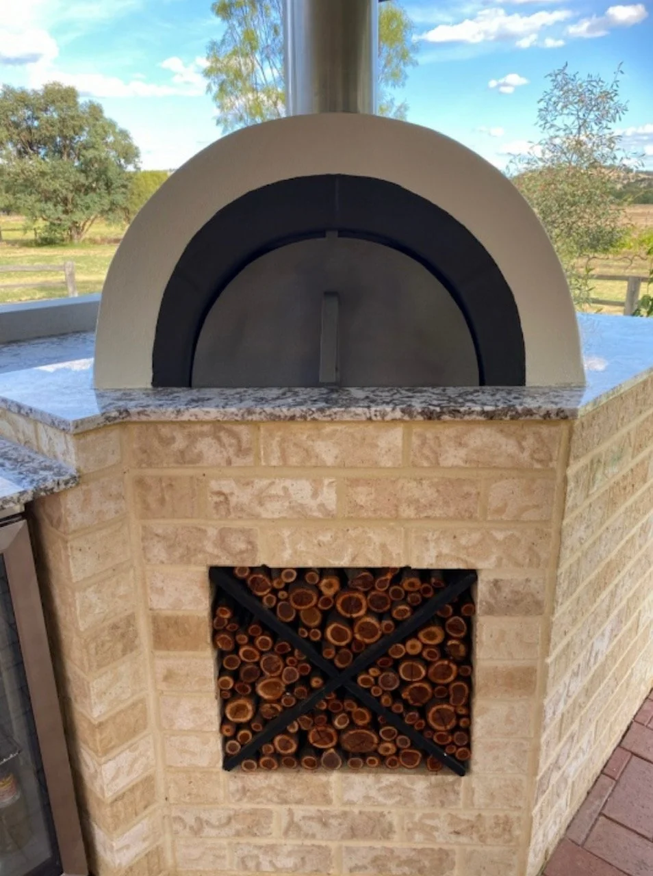 An outdoor pizza oven with a brick exterior, a granite countertop, and a firewood storage compartment filled with chopped logs.