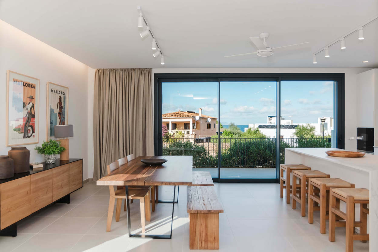 Interior of a modern living space with a large glass sliding door overlooking a balcony and trees outside, featuring a dining table with benches, bar stools, artwork on the walls, and various decorative objects.