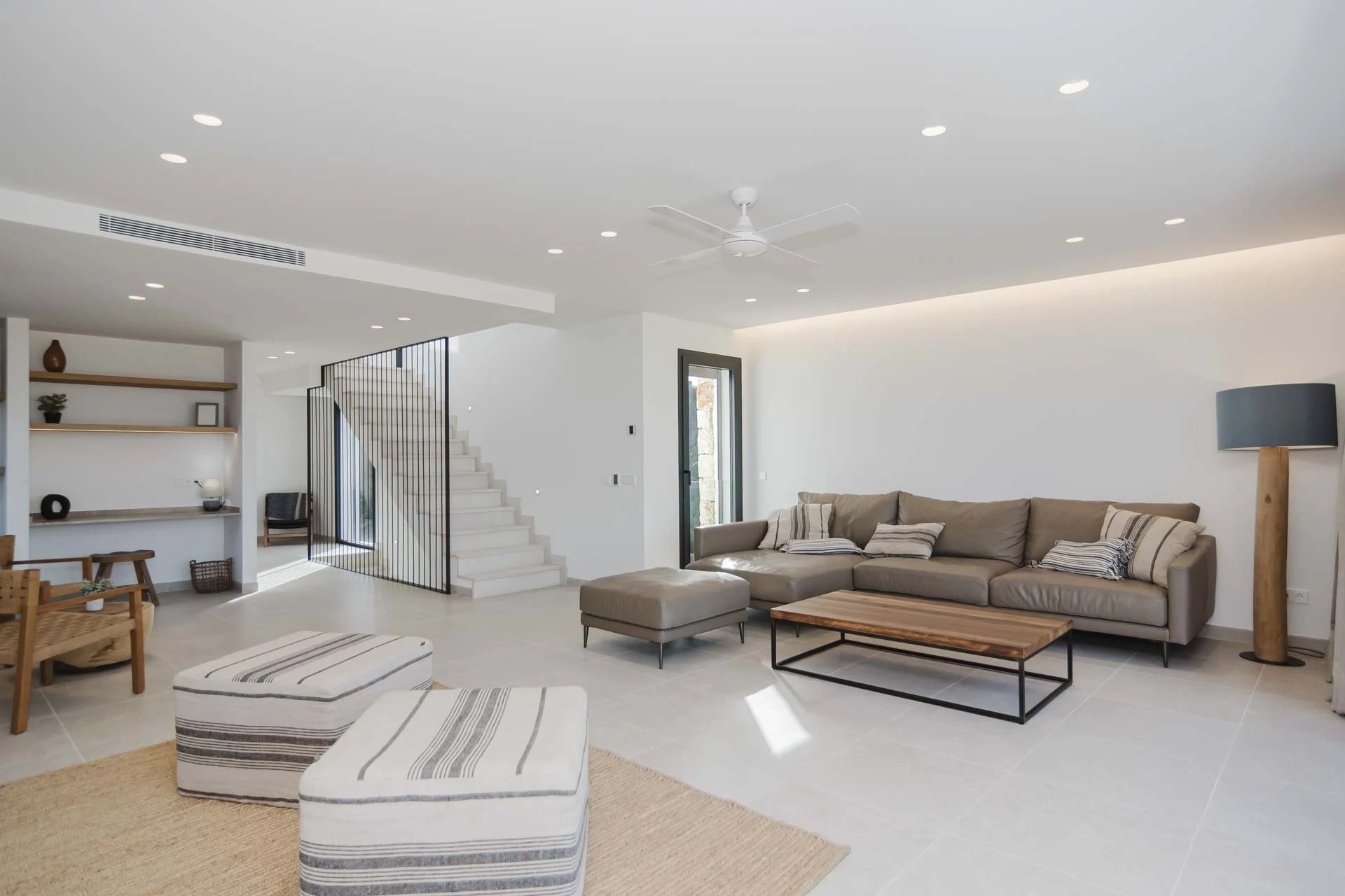 Modern living room with beige sectional sofa, striped pillows, wooden coffee table, floor lamp, white tiled flooring, and a staircase with black metal railing, bright natural light, and minimal decor.