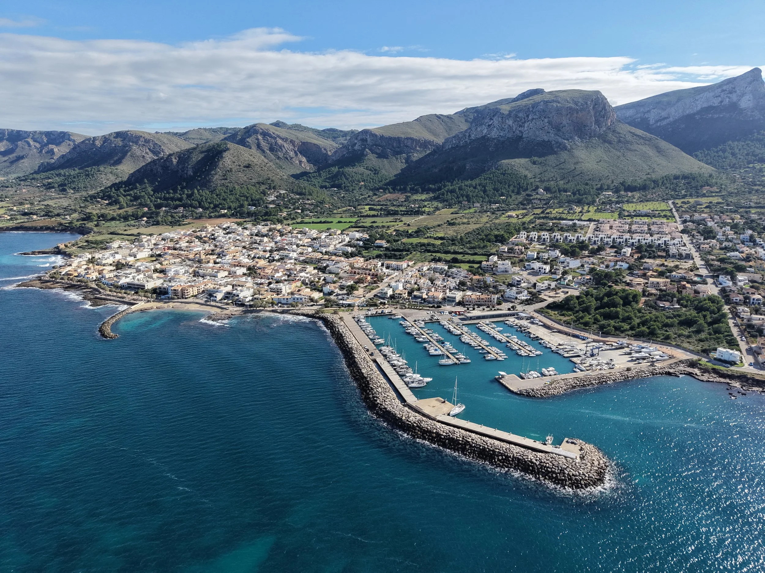 Aerial view of a seaside town with a marina, surrounded by mountains and a cloudy sky.