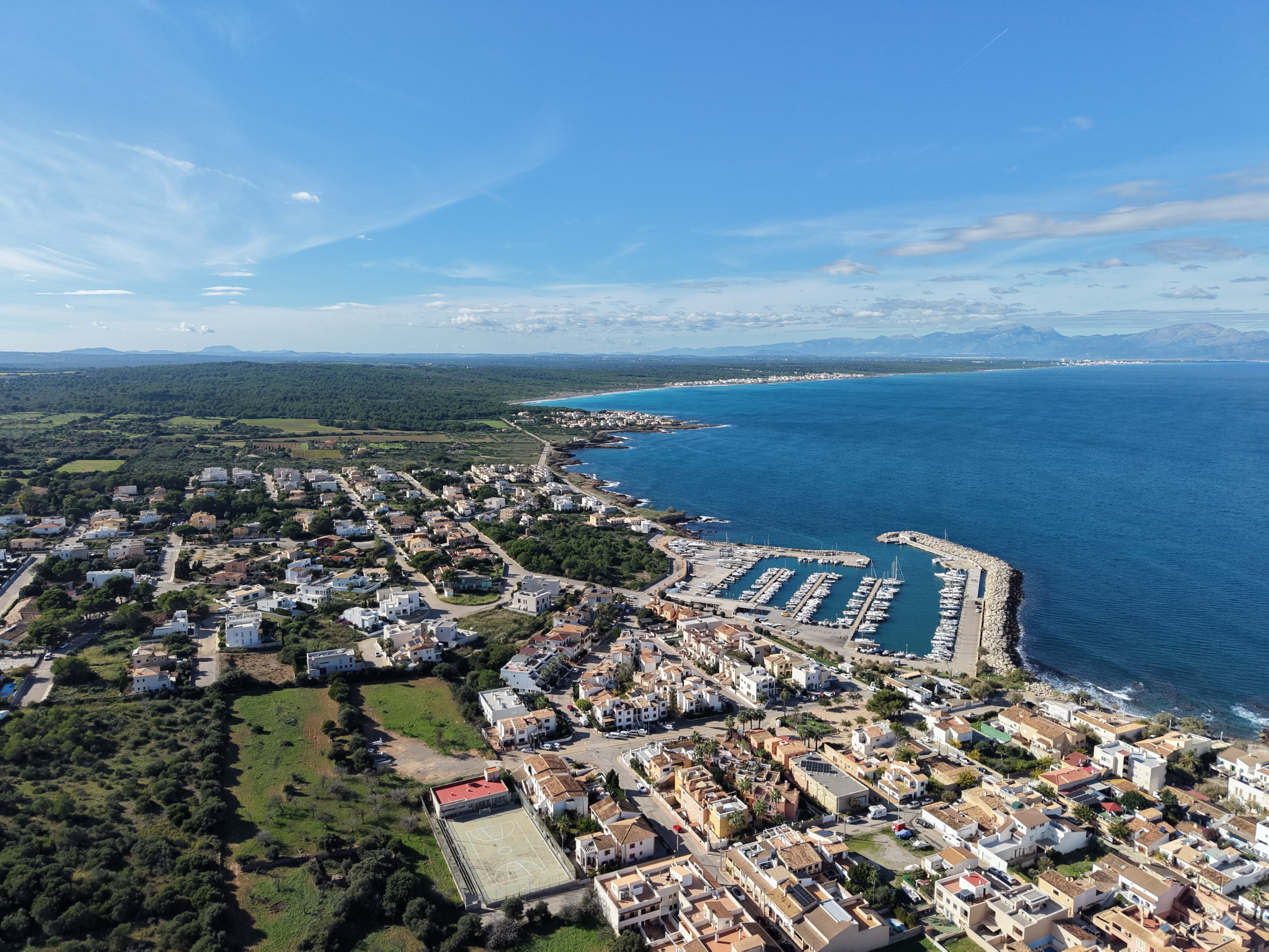 Aerial view of a coastal town with a marina, houses, and a basketball court near the shoreline under a blue sky.