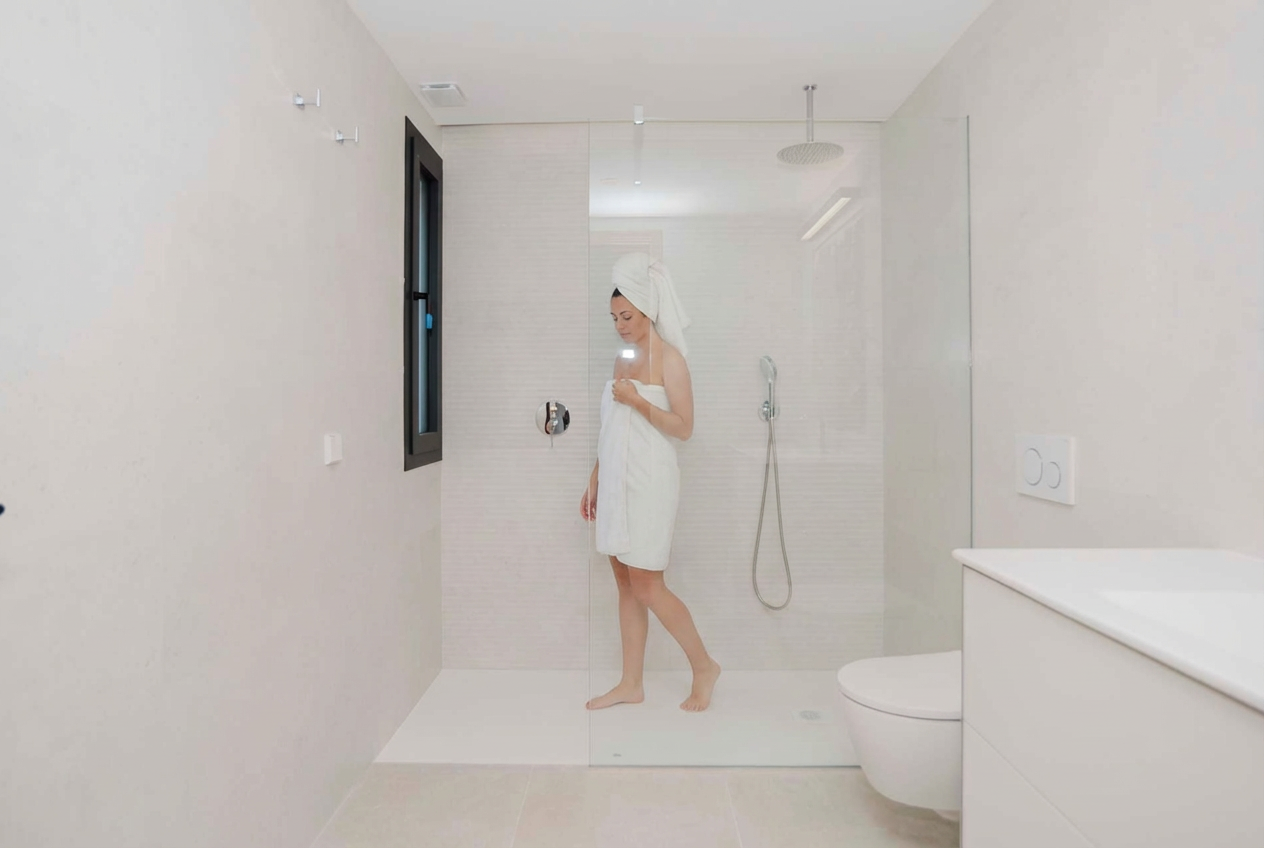 Woman wrapped in towel and with towel on her head, standing in a minimalist bathroom shower area, looking at her phone.