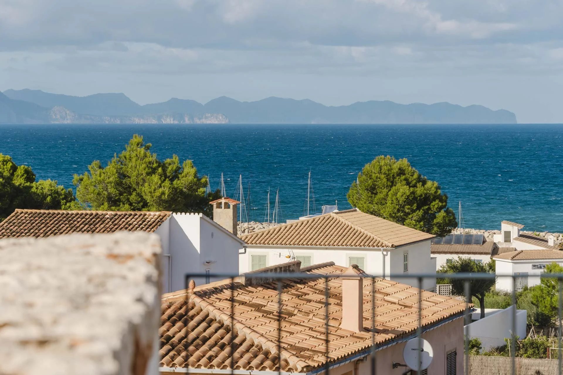 View of rooftops with orange tiles, green trees, the blue sea, and distant mountains under a partly cloudy sky.