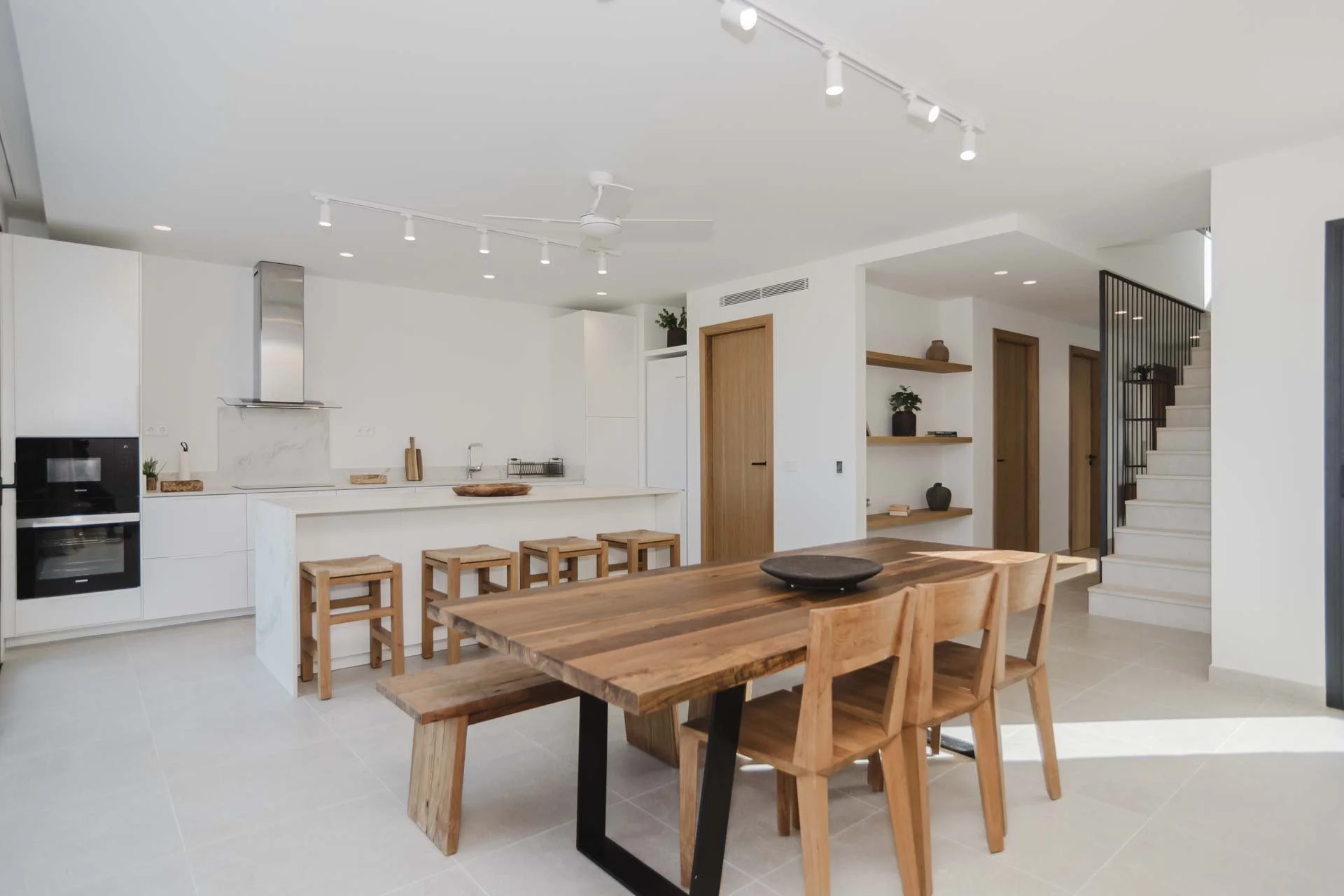 Modern open-plan kitchen and dining area with white cabinets, a wooden dining table, and wooden bar stools. There are shelves with decorative items, a staircase with black railing, and light-colored tiled floors.