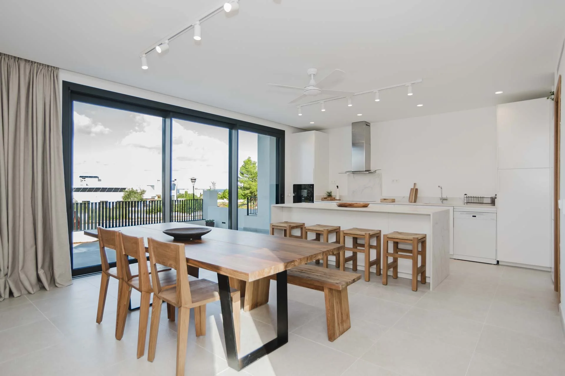 Modern open-concept kitchen and dining area with large sliding glass doors leading to a balcony, featuring a wooden dining table with chairs, a kitchen island with stools, white cabinets, and a stainless steel range hood.