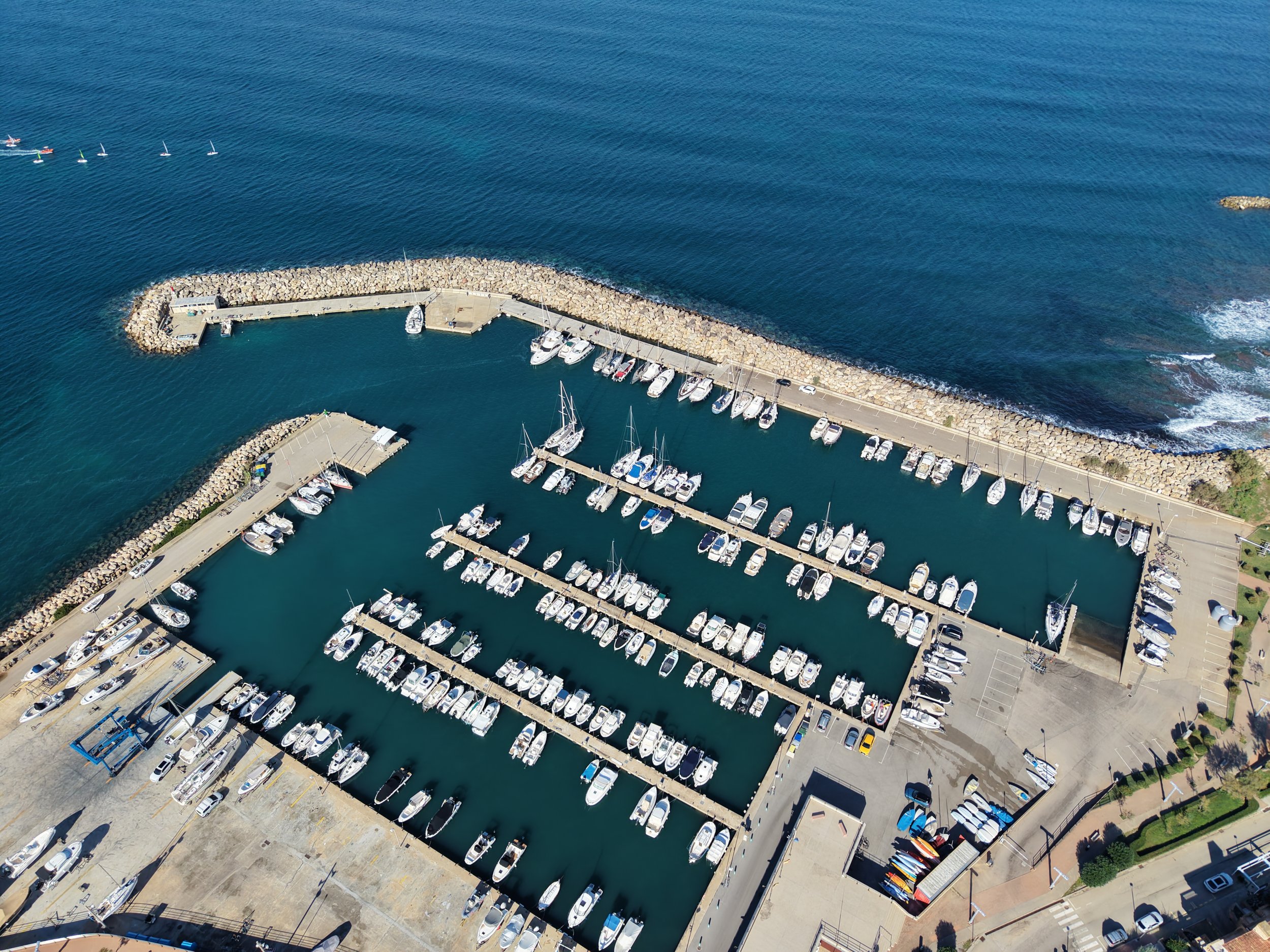 Aerial view of a harbor with boats docked along multiple piers, surrounded by a rocky breakwater and open ocean.