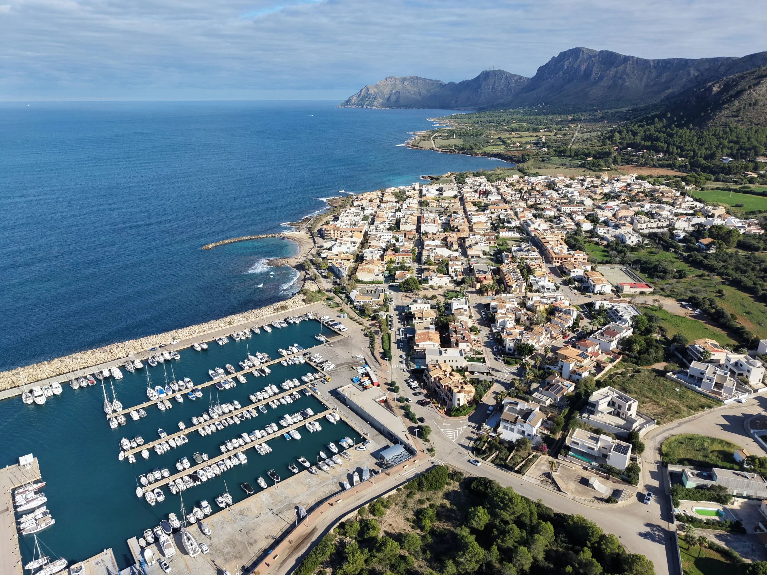 Aerial view of a coastal town with a marina filled with boats, a rocky breakwater, and a scenic backdrop of mountains and the ocean.