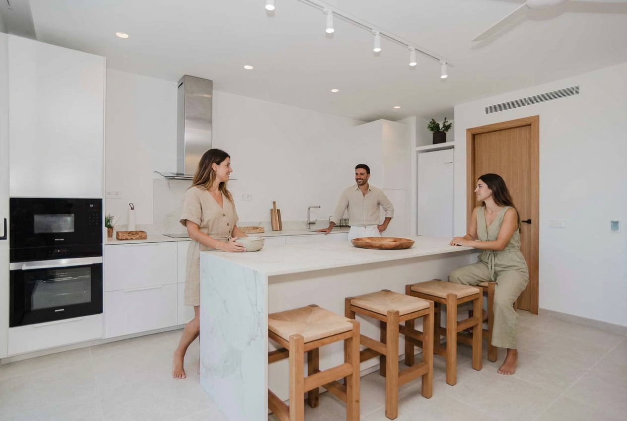 Three people in a modern white kitchen. Two women and one man are sitting and standing around a marble island counter, engaging in conversation. The kitchen has built-in appliances and simple decor.