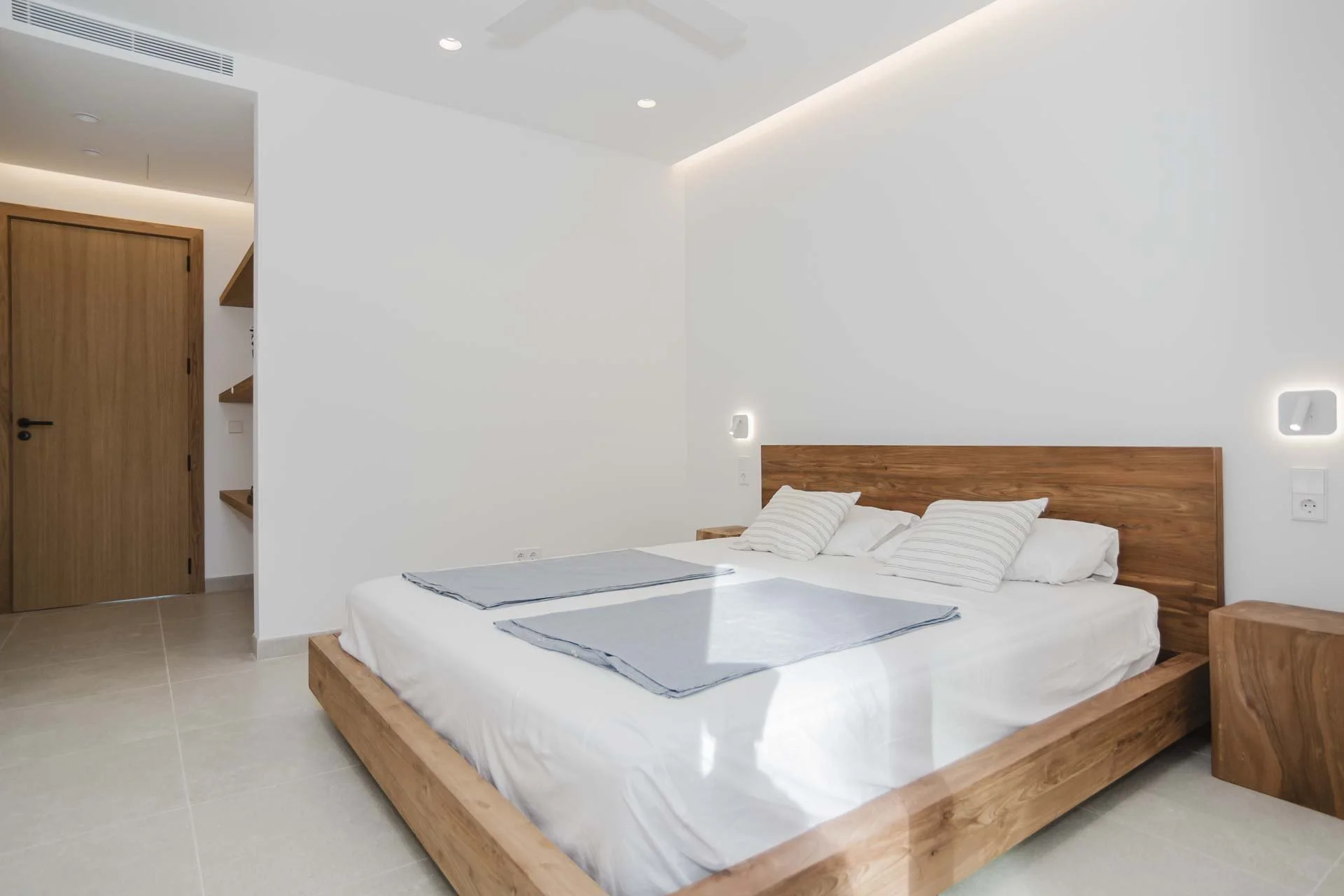 Modern minimalist bedroom with a wooden bed frame, white bedding, and striped pillows. Bare white wall with subtle lighting, wooden door, and small corner shelves.