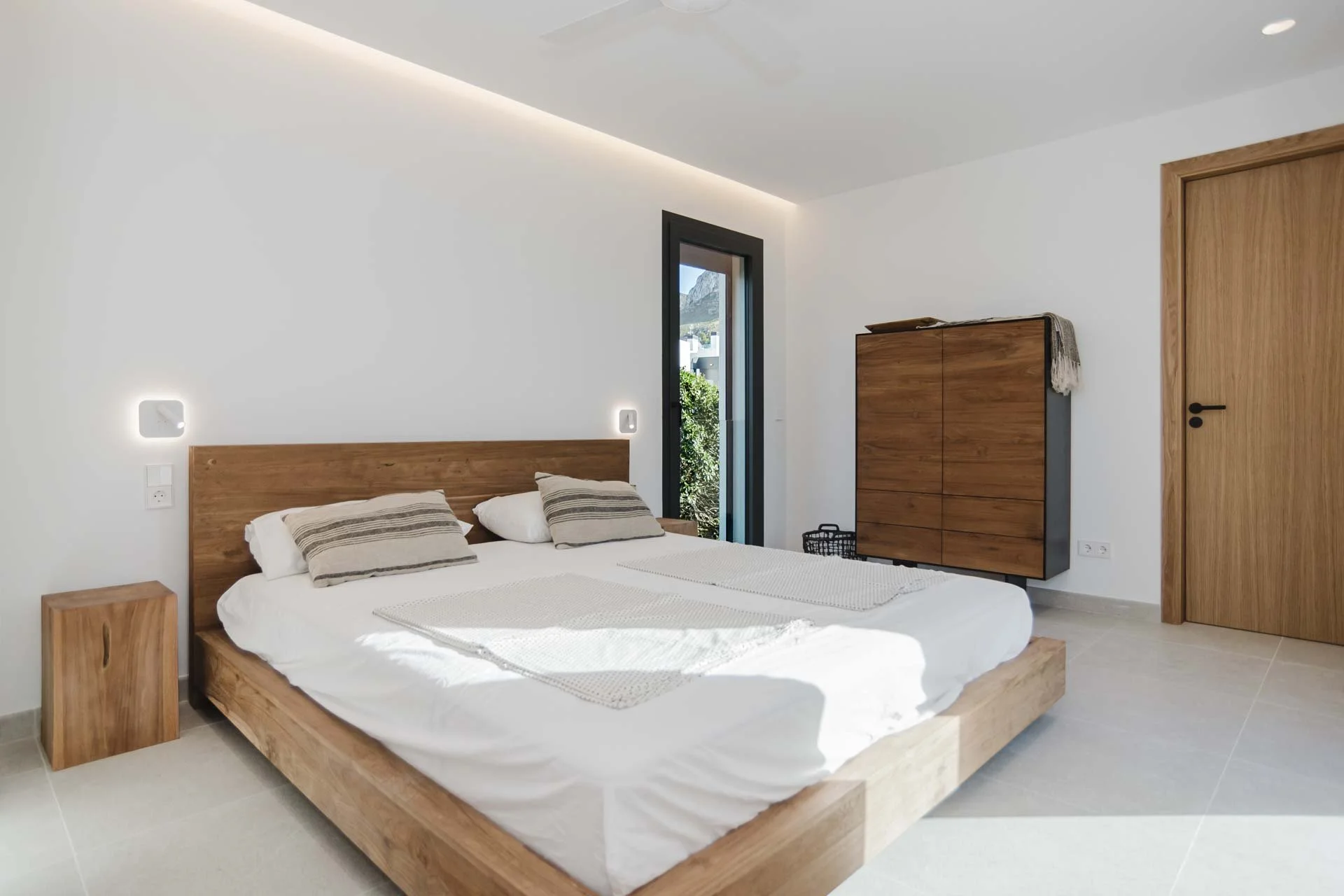 Minimalist bedroom with wooden furniture, white walls, and large bed with beige striped pillows, sunlight streaming in through a door, and a wooden wardrobe.