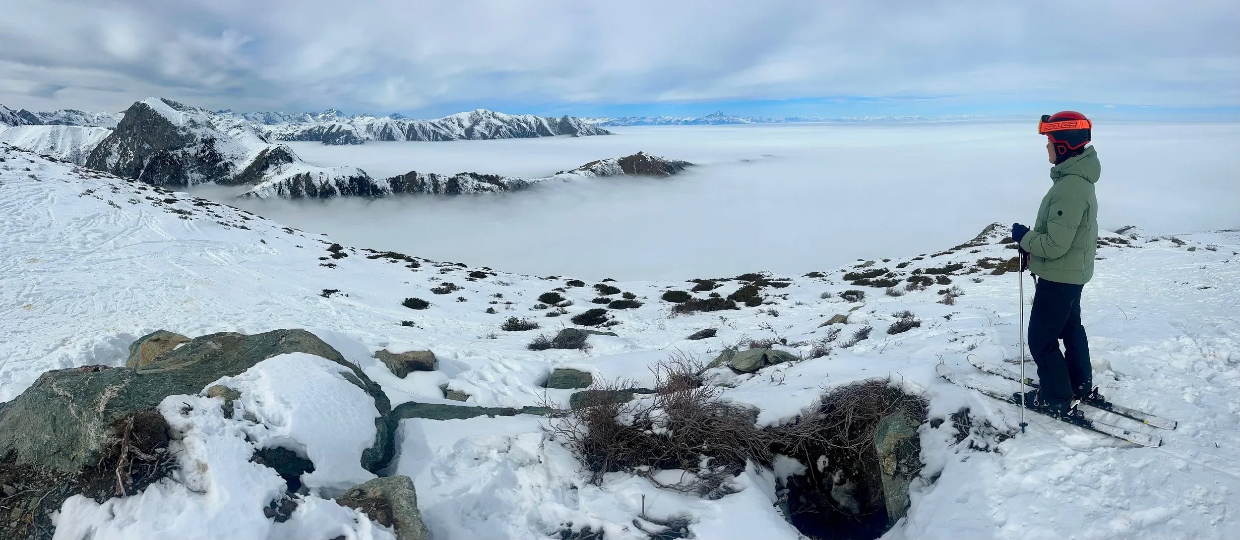A person wearing a green jacket, black pants, and a red ski helmet stands on snow-covered ground with skis and poles, overlooking a snowy mountain landscape with fog and clouds below and snow-capped mountains in the distance.