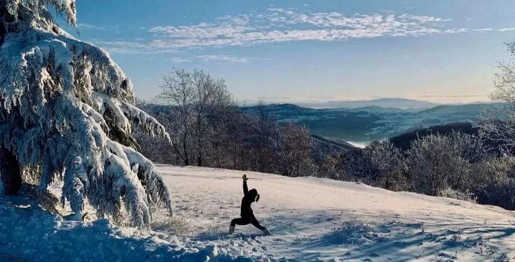 A person practicing yoga on a snow-covered hillside with snowy trees and distant mountains in the background under a partly cloudy sky.