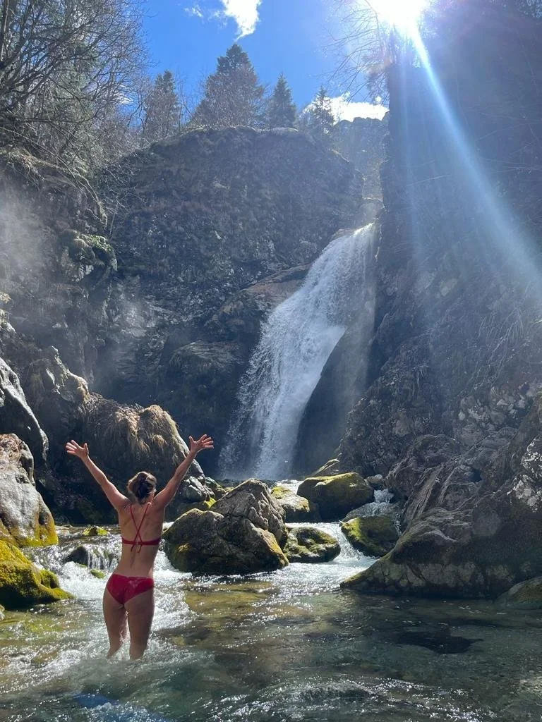 Woman in red bikini standing in a creek with arms raised, near a waterfall in a rocky, wooded area on a sunny day.
