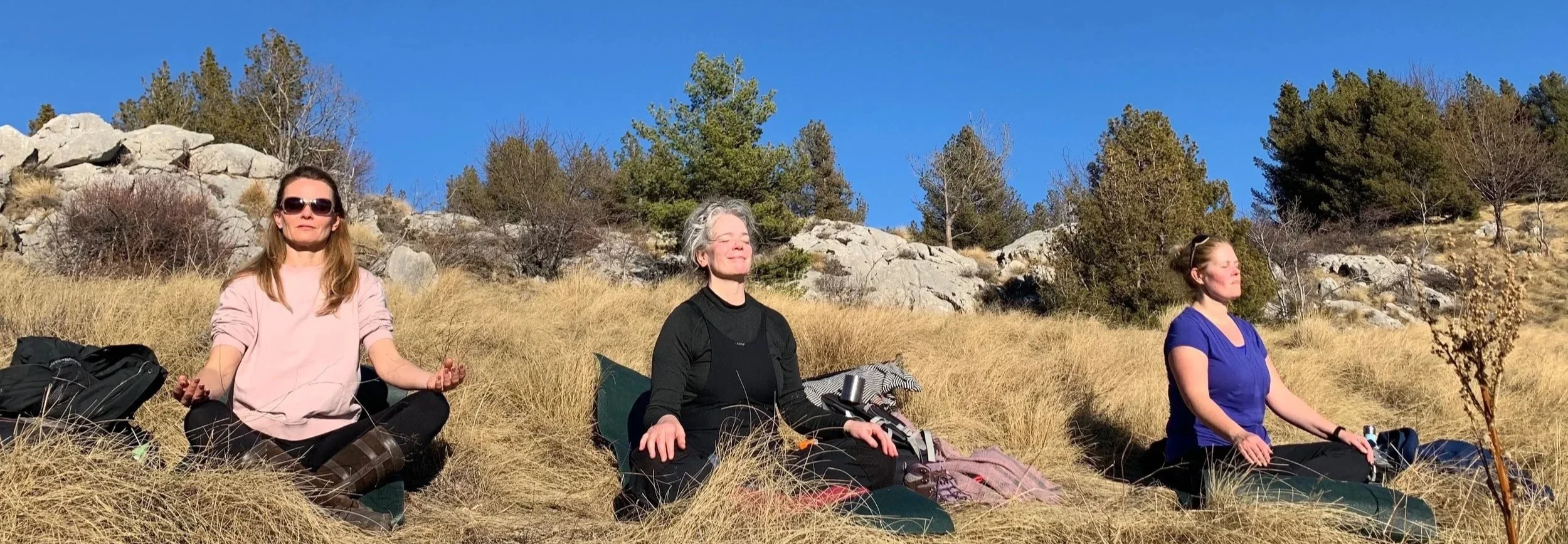 Three women sitting cross-legged on the grass with their eyes closed, meditating outdoors on a sunny day, surrounded by rocks, trees, and dry grass.