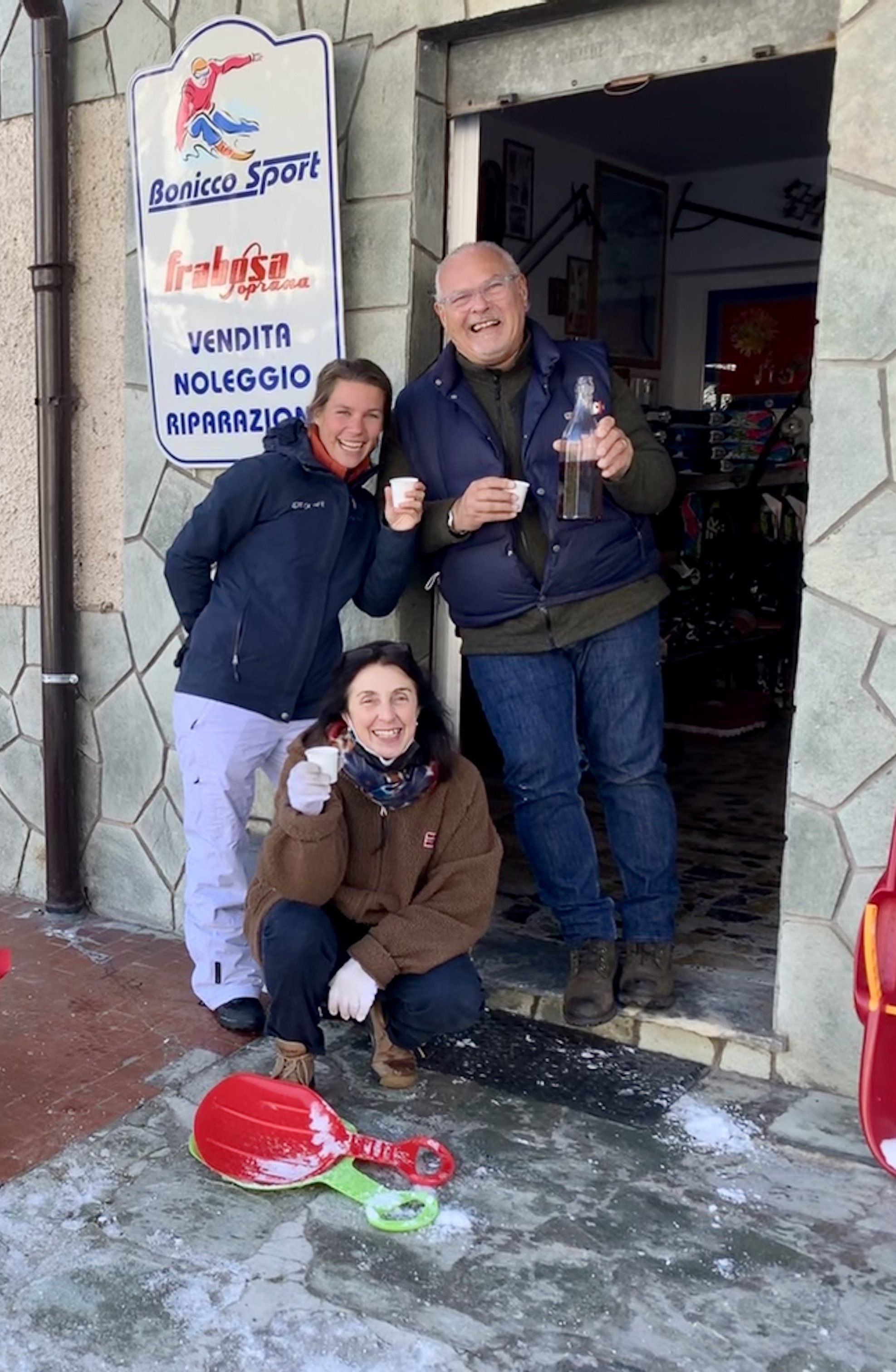 Three people smiling and holding drinks outside an ice skating rink, with a sign that reads 'Bonico Sport' and 'Vendita Noleggio Riparazione.' There is a red and green plastic snow shovel on the ground and a red sled.