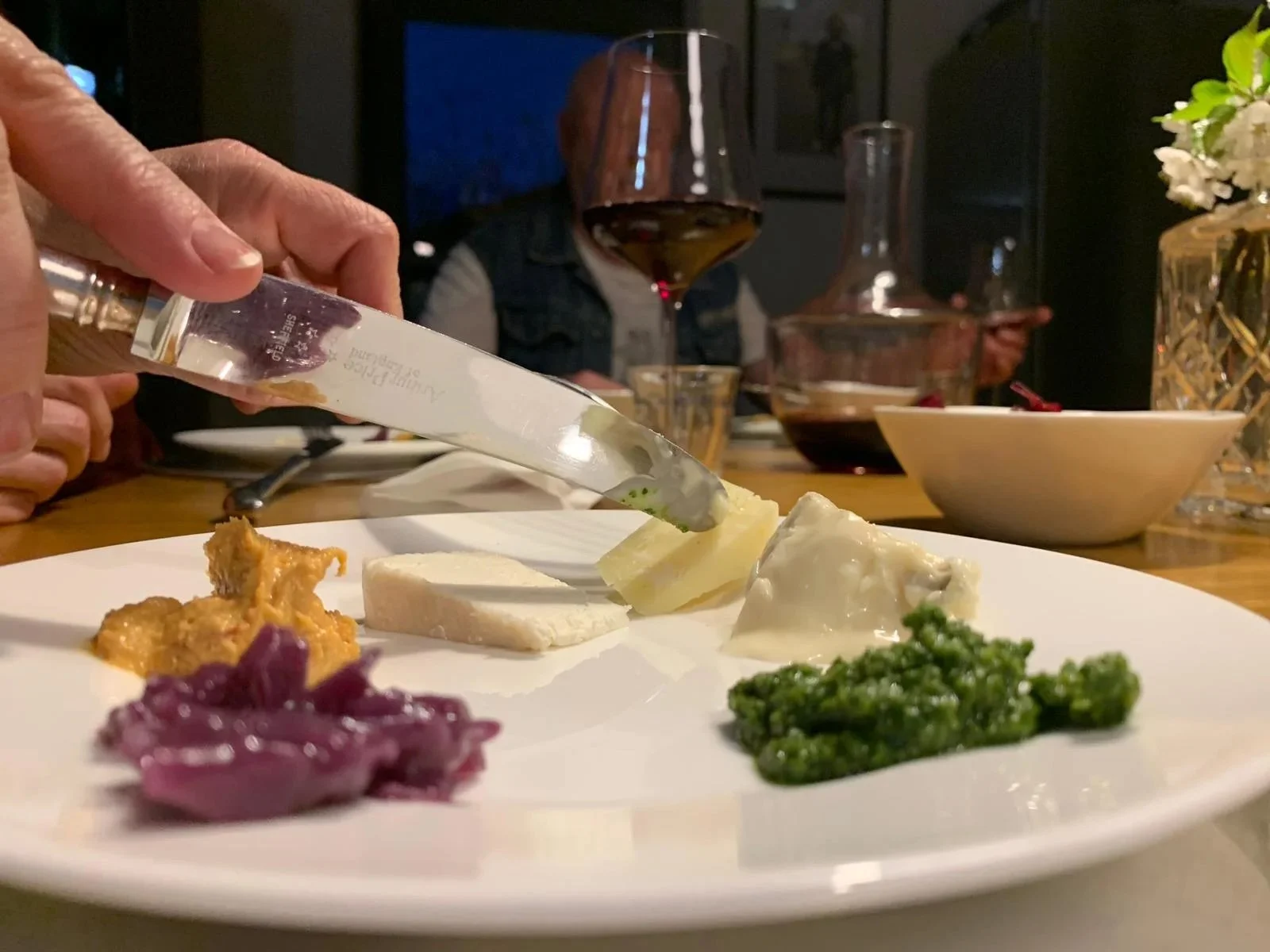 Person cutting cheese on a white plate with assorted cheeses, pickled vegetables, and greens, at a dinner gathering with wine and glasses in the background.