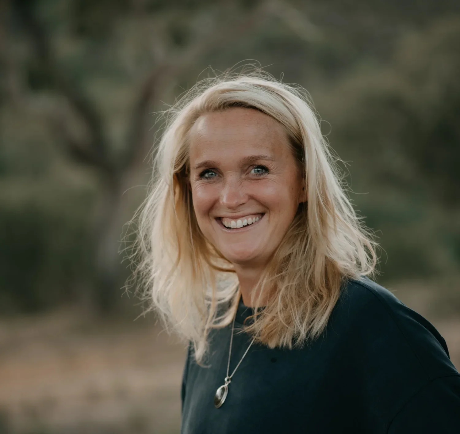 A woman with blonde hair and blue eyes smiling outdoors, wearing a black top and a silver necklace.