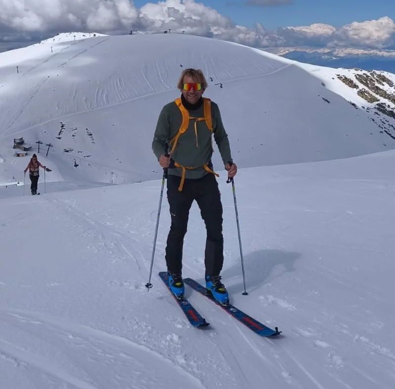 A man in gray and black ski gear with yellow accents and reflective sunglasses stands on skis in a snowy mountain landscape, holding ski poles, with another skier in the background.
