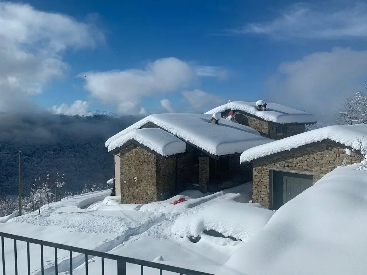 Snow-covered stone house with multiple roofs blanketed in snow, hills and cloudy sky in the background.