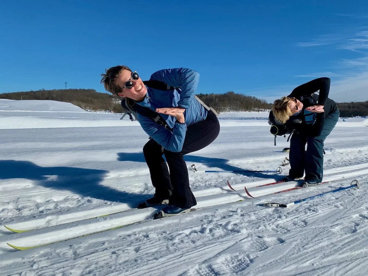 Two people in winter gear on skis in a snowy landscape, posing playfully and smiling at the camera, with a blue sky and distant hill in the background.