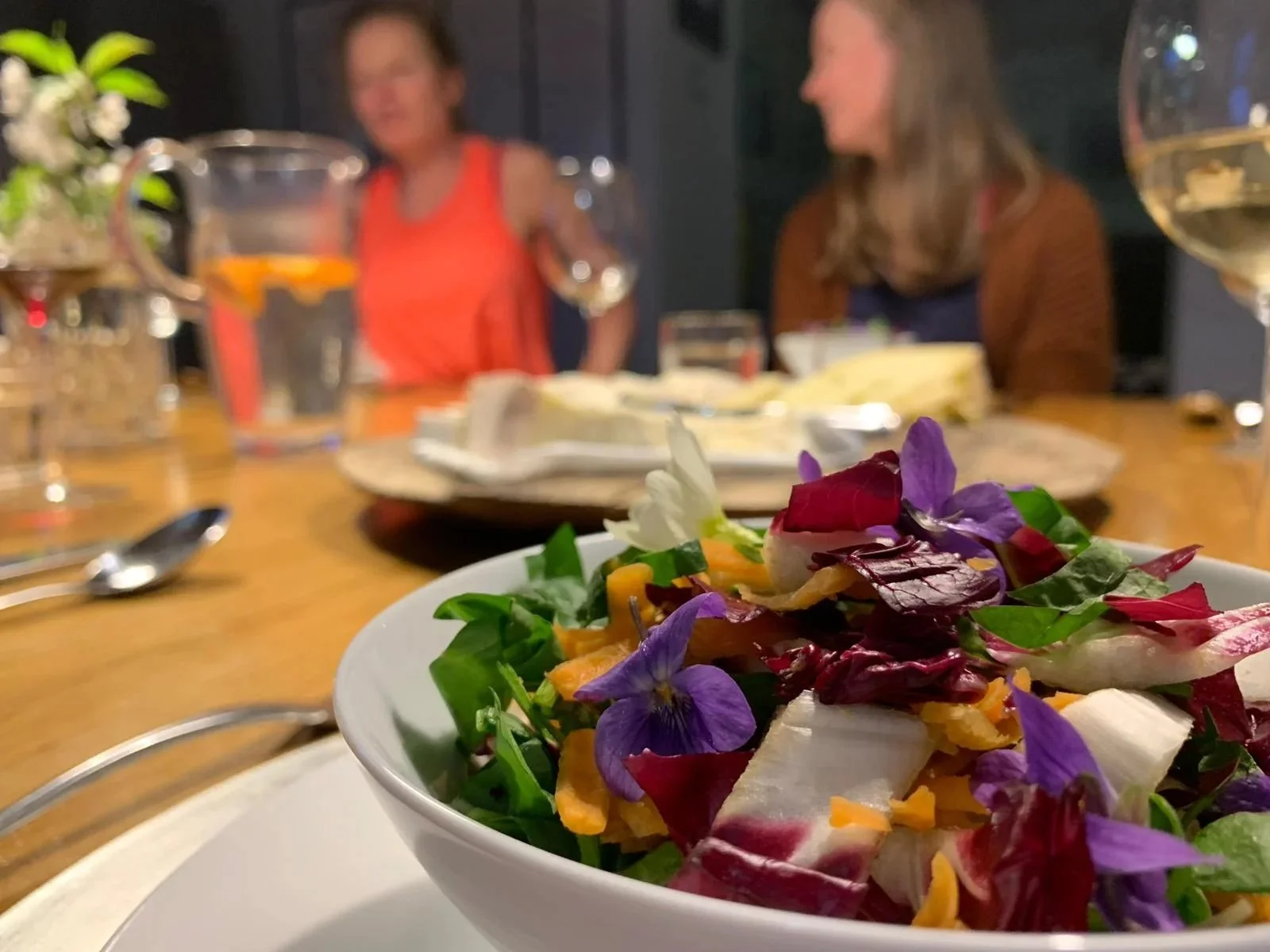 Close-up of a salad with colorful greens, flowers, and shredded cheese, on a white plate at a dinner table with two blurred women in the background, engaged in conversation.