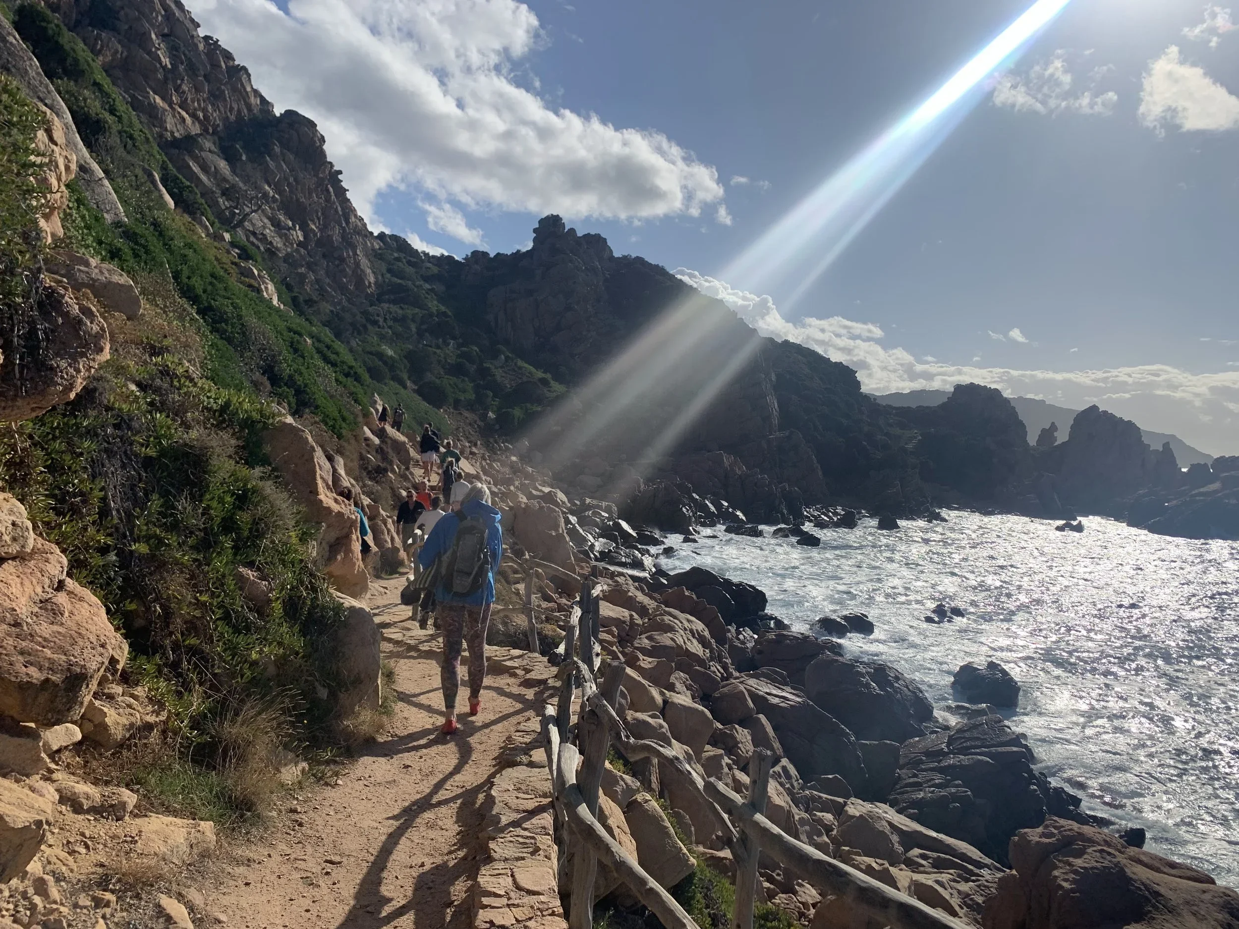 A group of people hiking along a rocky coastal trail with a wooden fence on the right, next to the ocean with waves, rocky cliffs and a mountain range in the background, under a partly cloudy sky with a bright sunbeam.