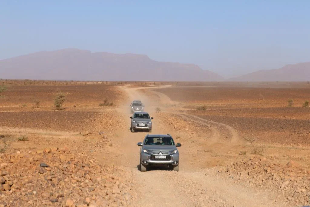 Four SUVs driving on a dusty, rocky desert road with mountains in the distance under a clear blue sky.