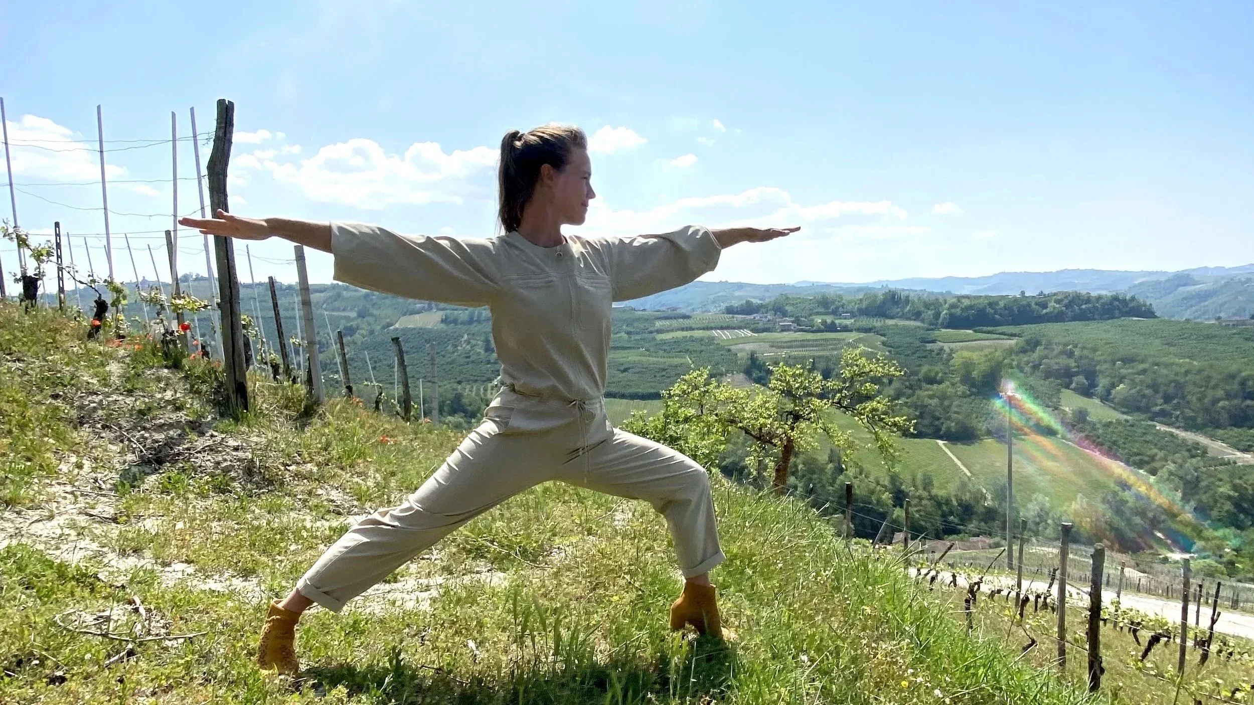 A woman practicing yoga in a wide-open field overlooking a landscape of rolling hills and vineyards on a sunny day.