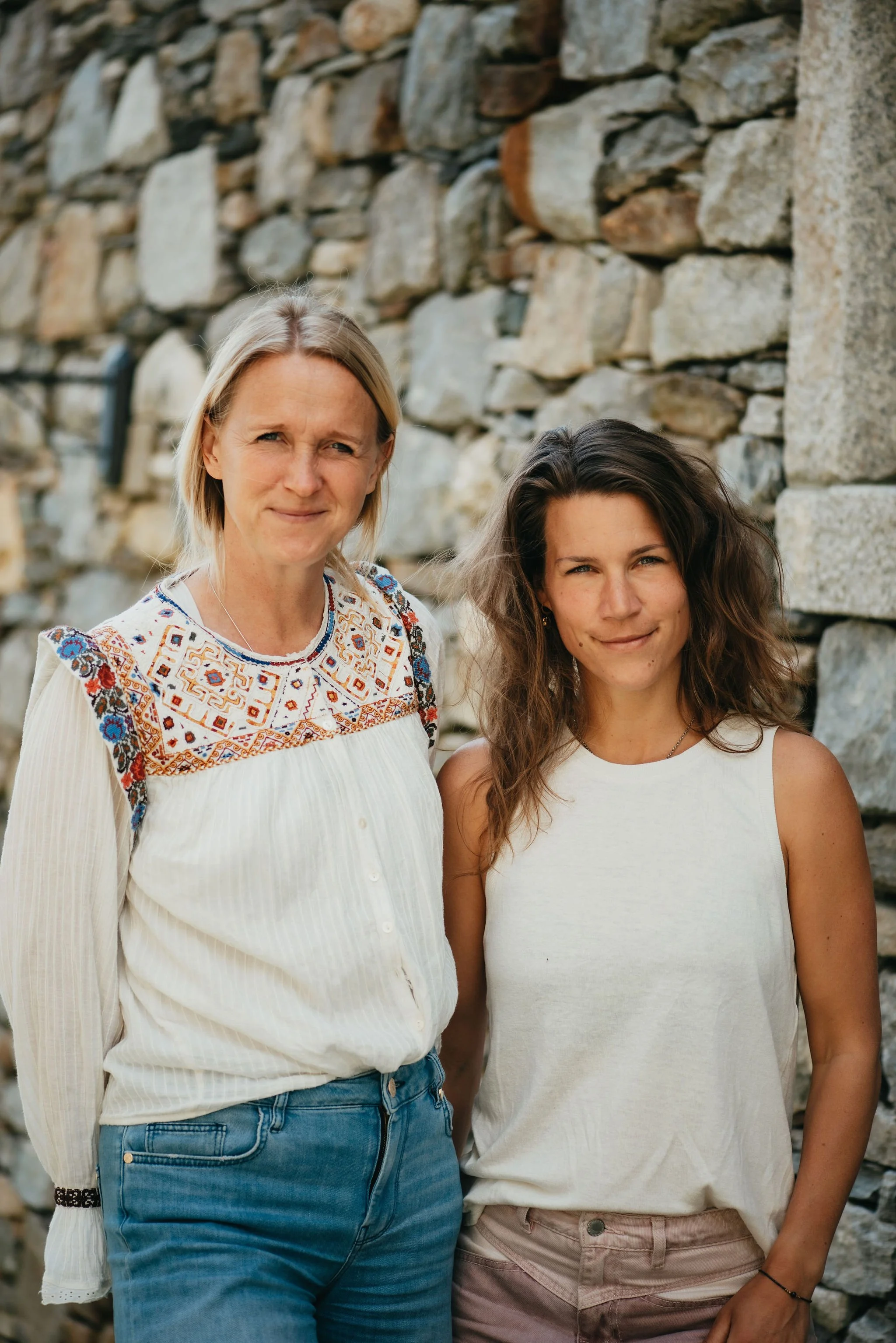 Two women standing outdoors in front of a stone wall, smiling at the camera.