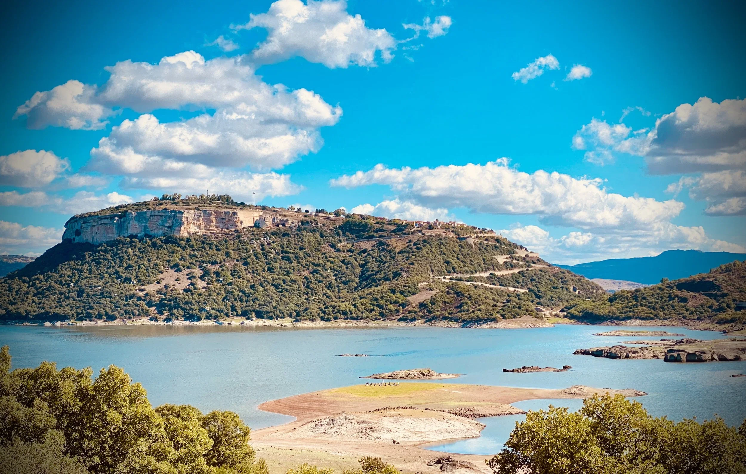 Landscape view of a large reservoir or lake surrounded by green trees and hills under a partly cloudy sky.