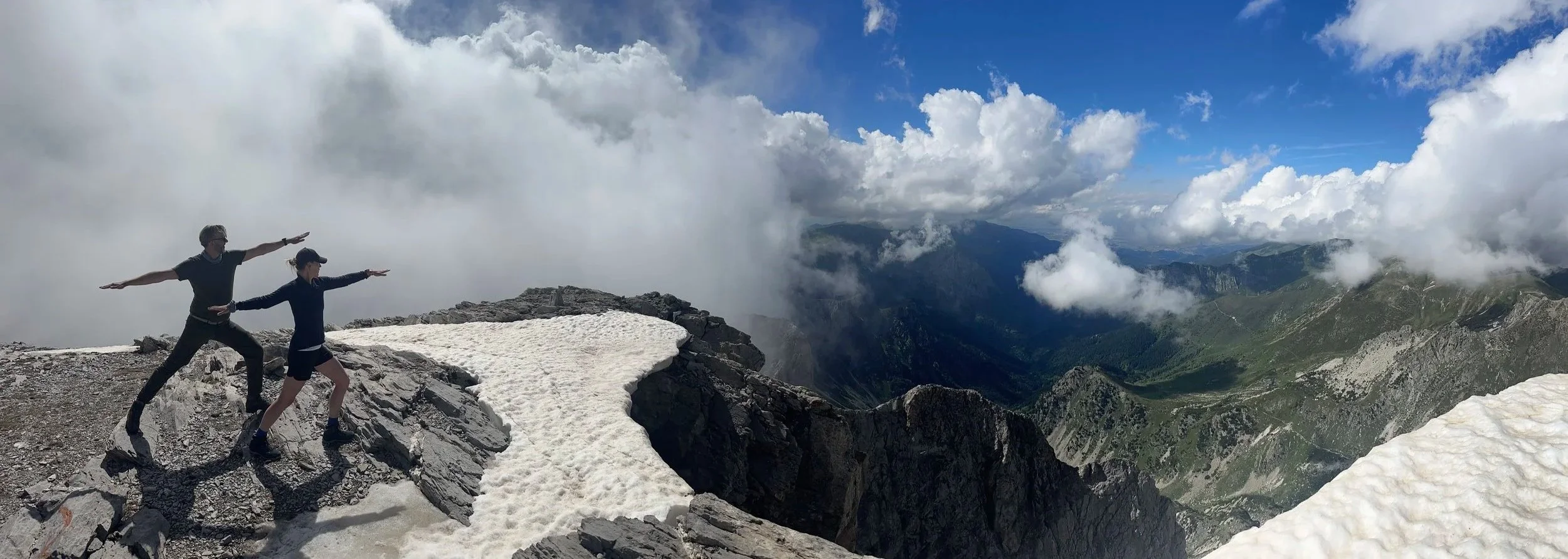 Two hikers standing on a mountain ridge with snow patches, arms outstretched, overlooking a mountain range with clouds and blue sky.