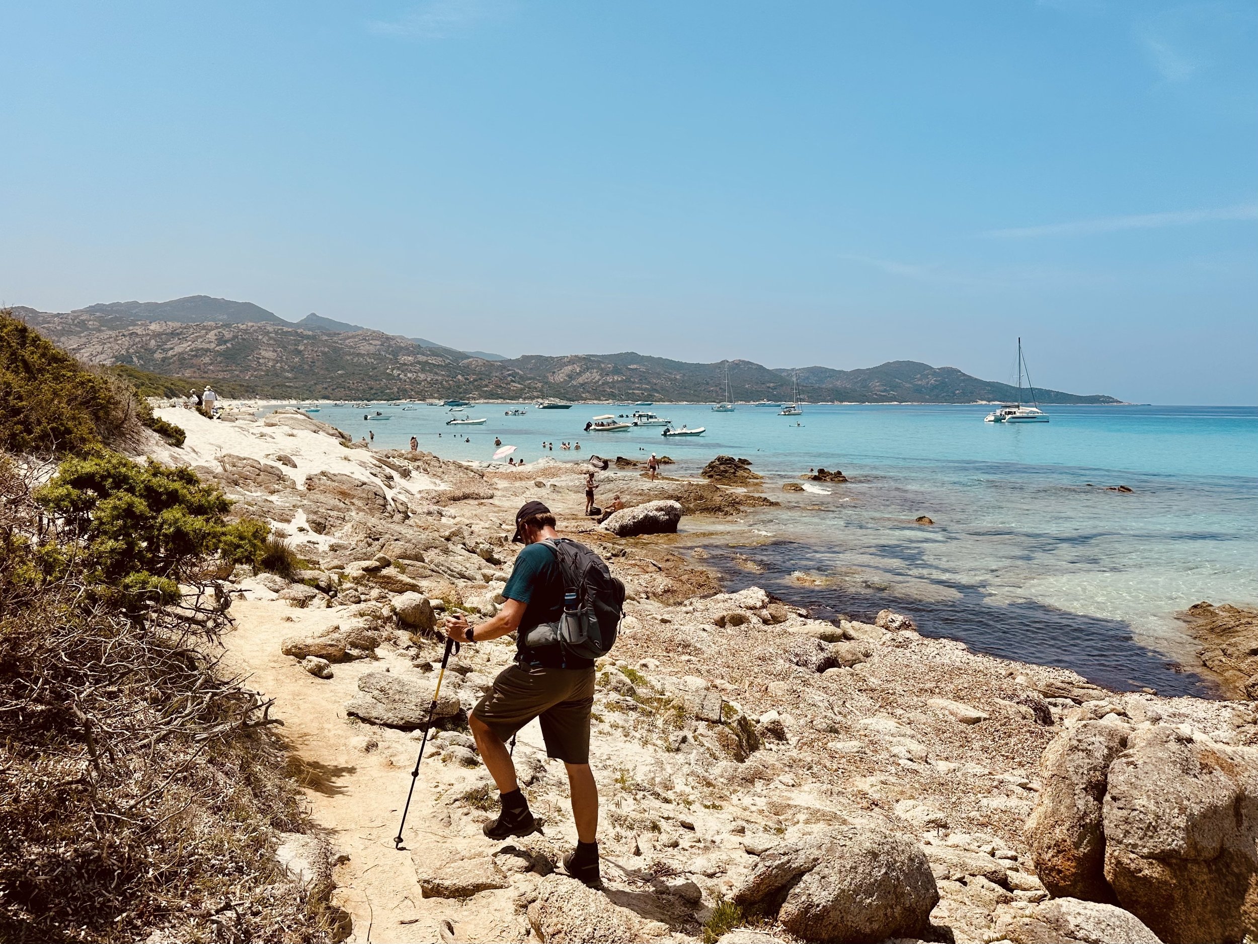 Hiker climbing rocky terrain on a beach with turquoise water and sailboats in the distance under a clear sky.