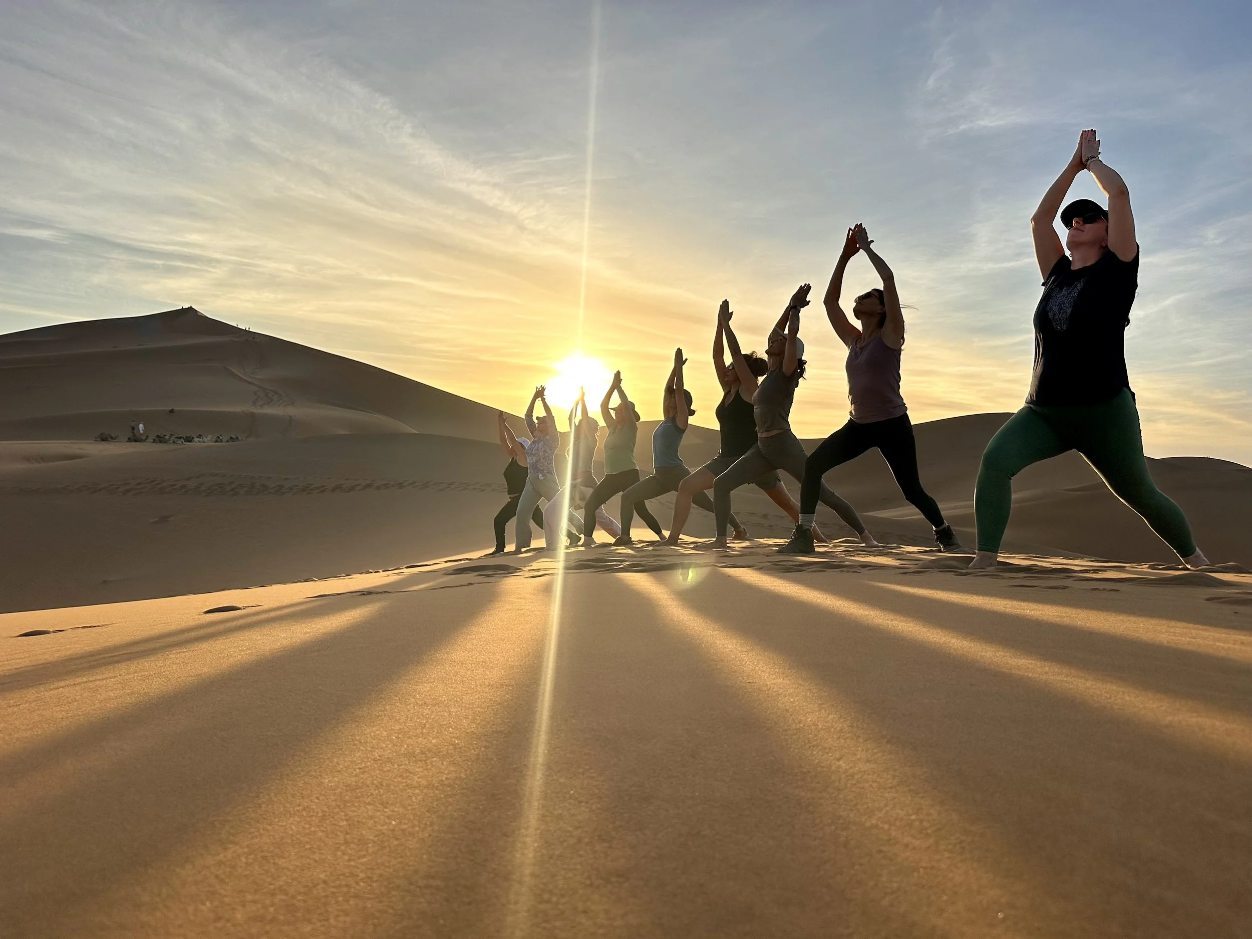 A group of people practicing yoga in a desert at sunset, standing on the sand with their hands raised above their heads.