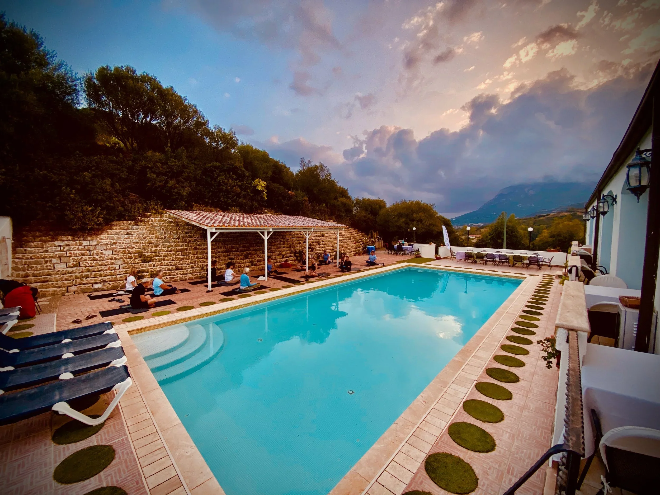 People practicing yoga beside a swimming pool during sunset at a mountain resort.