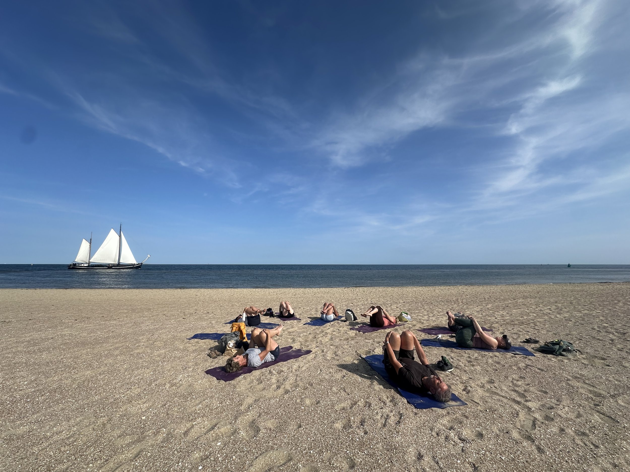 People practicing yoga on mats on a sandy beach near the ocean with a sailboat in the distance and a partly cloudy blue sky.