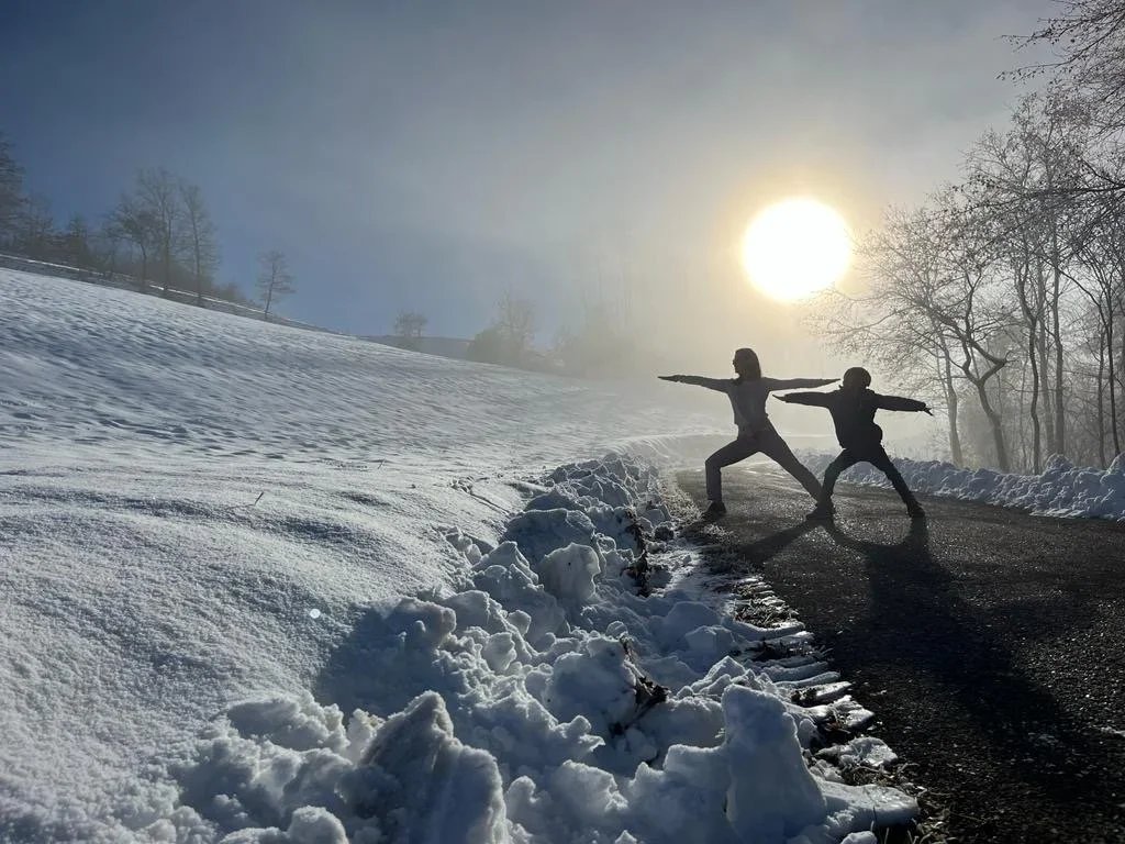 Two people in athletic clothing perform yoga poses with arms extended on a winding mountain road during winter, with snow-covered ground, bare trees, and a low sun in the background.