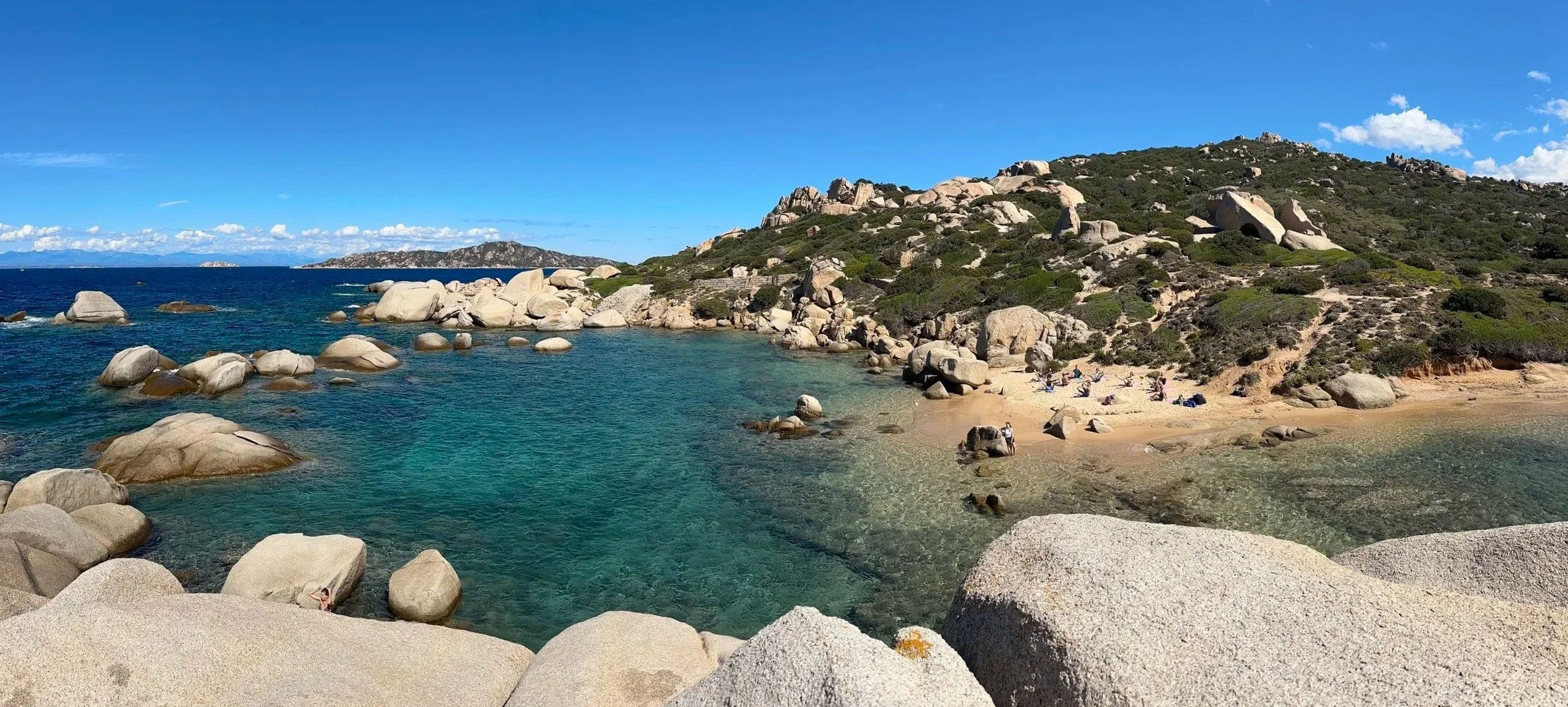 A rocky coastal landscape with clear turquoise water, a sandy beach, large boulders, and a hillside covered in green shrubs and rocks under a blue sky with some clouds.