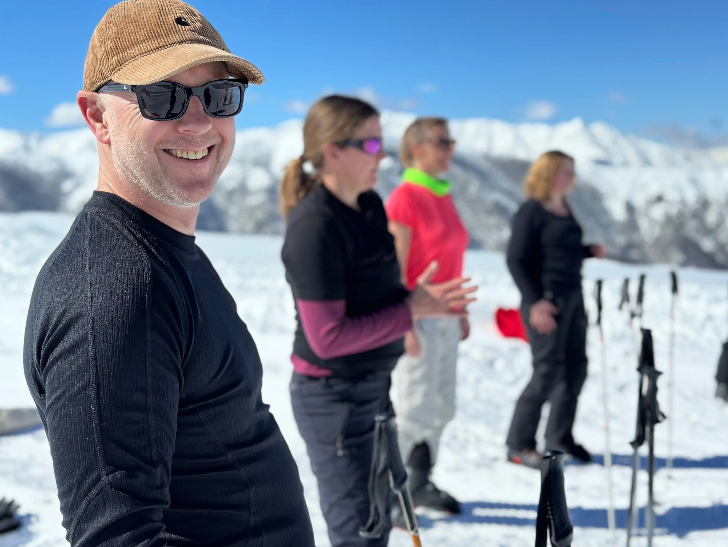 Group of people skiing in a snowy mountain landscape, with mountains in the background, some wearing sunglasses and winter sports gear.