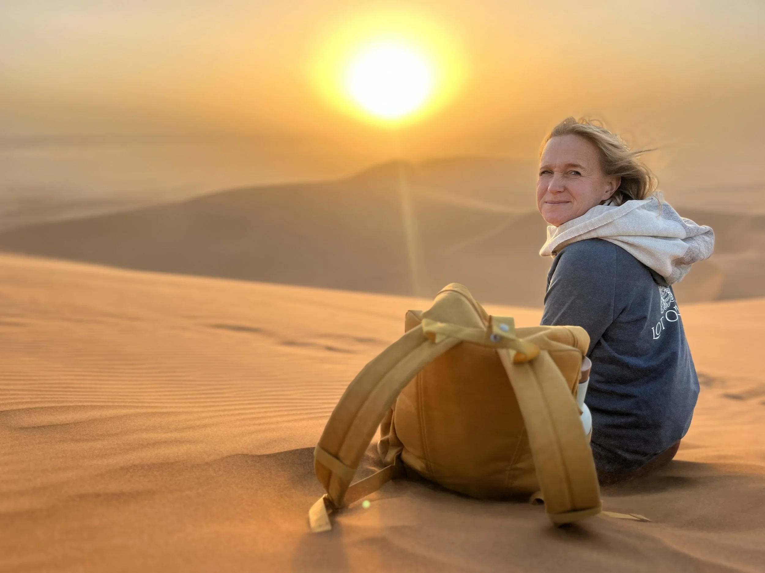 A woman sitting on sand dunes during sunset with a yellow backpack beside her.