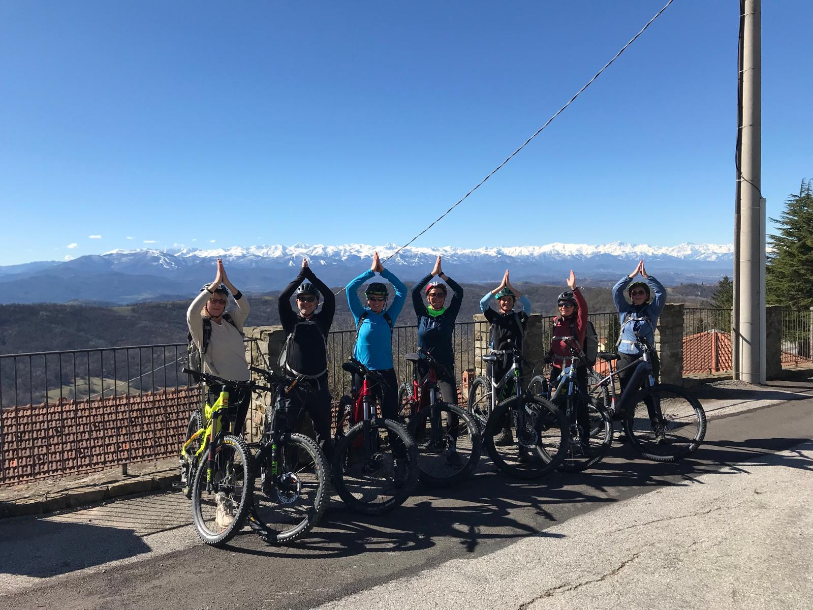 Group of seven cyclists standing with their bikes on a mountain overlook, making a prayer gesture with their hands above their heads, snow-capped mountains in the background on a clear day.