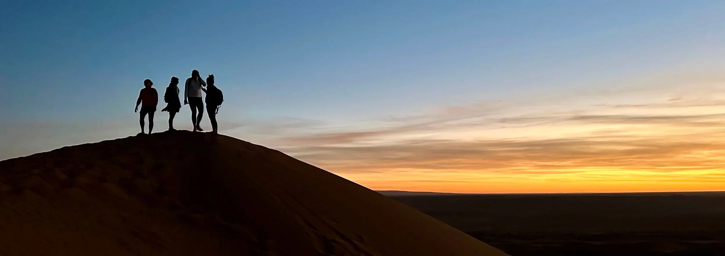 Silhouettes of four people standing on a sand dune at sunset in a desert landscape.