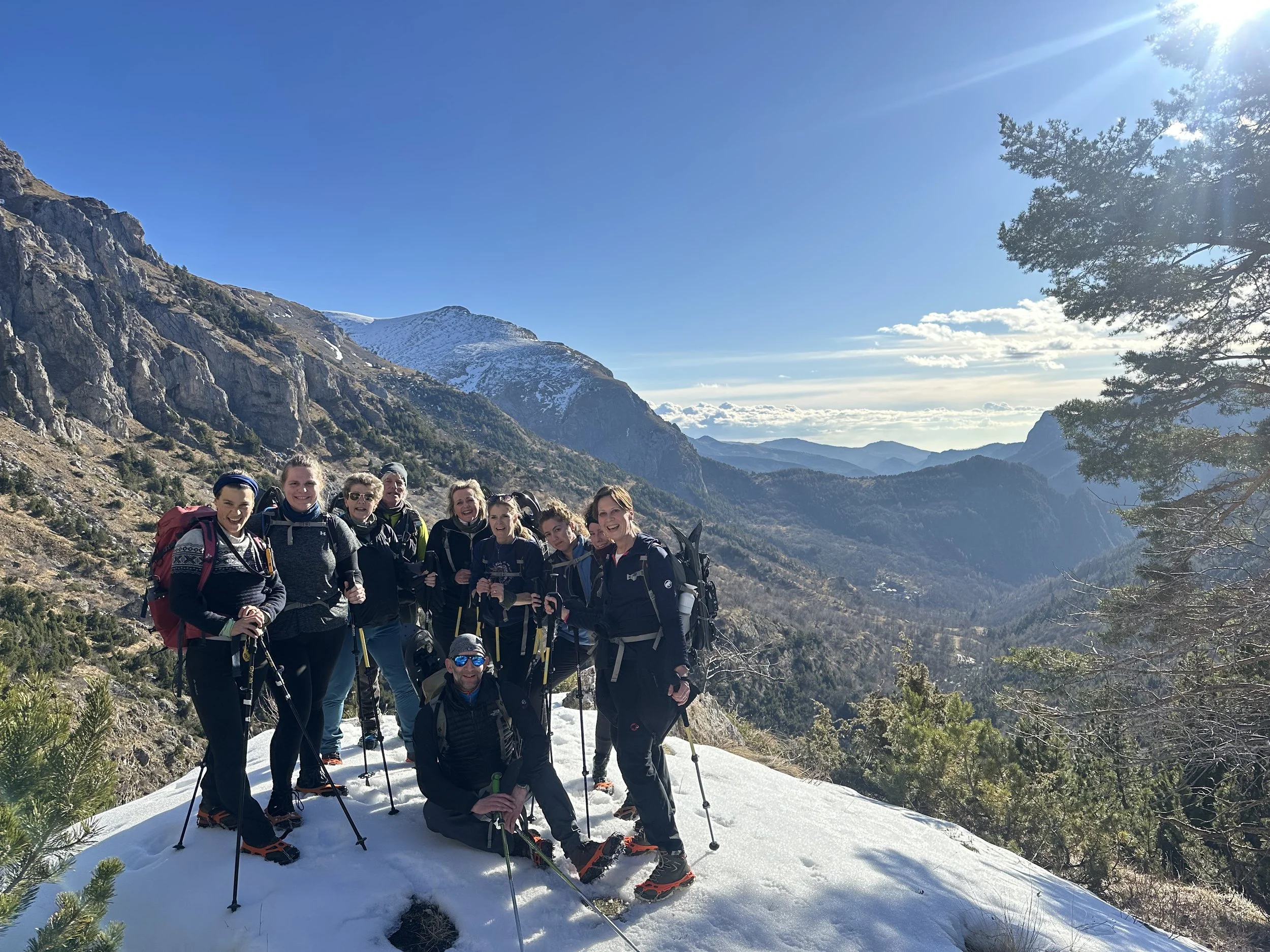 Group of hikers standing on snow-covered ground in a mountainous landscape with rocky cliffs, snow-capped peaks, and clear blue sky in the background.