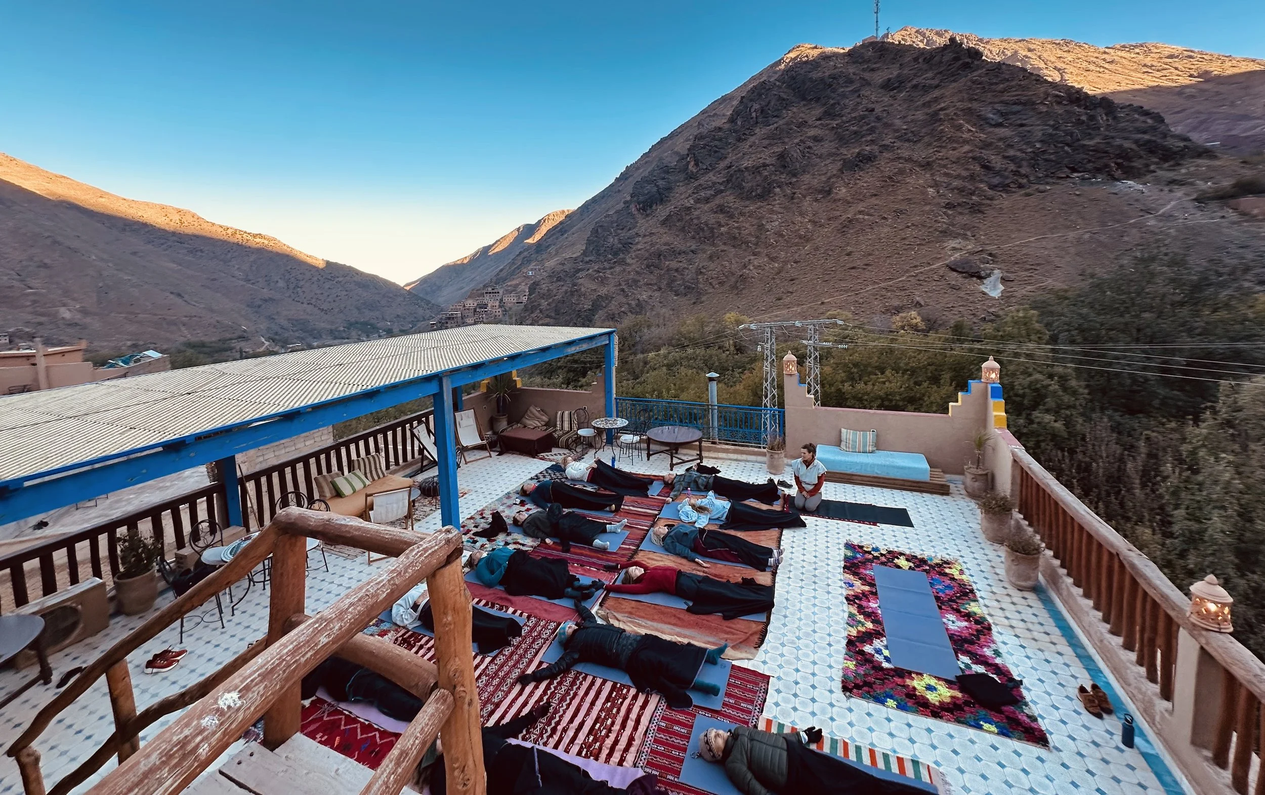 People practicing yoga or meditation on a rooftop terrace with colorful rugs and blankets, surrounded by mountains and clear sky