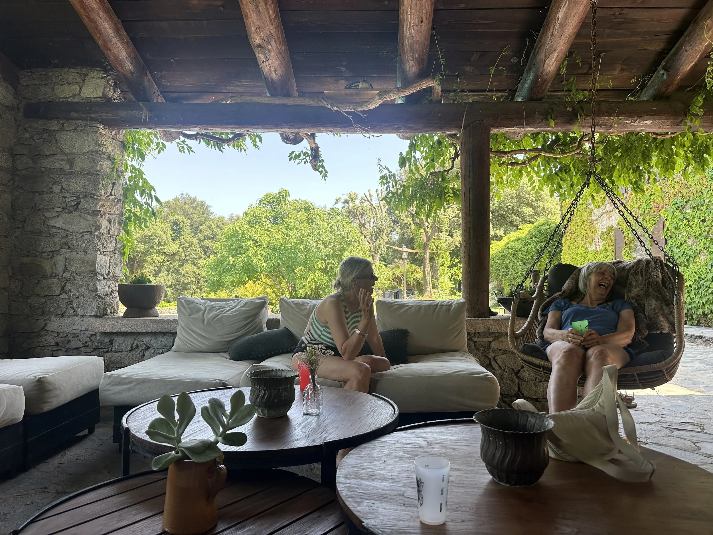 Two women are sitting and laughing in a cozy outdoor seating area with stone walls, wooden beams, and lush green trees in the background. One woman is reclining on a swinging chair holding a green cup, and the other is sitting on a cream-colored sofa with cushions.
