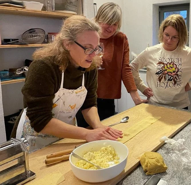 Three women in a kitchen preparing pasta dough, with one woman rolling out the dough and the other two observing, a bowl of grated cheese, and pasta-making tools on the counter.
