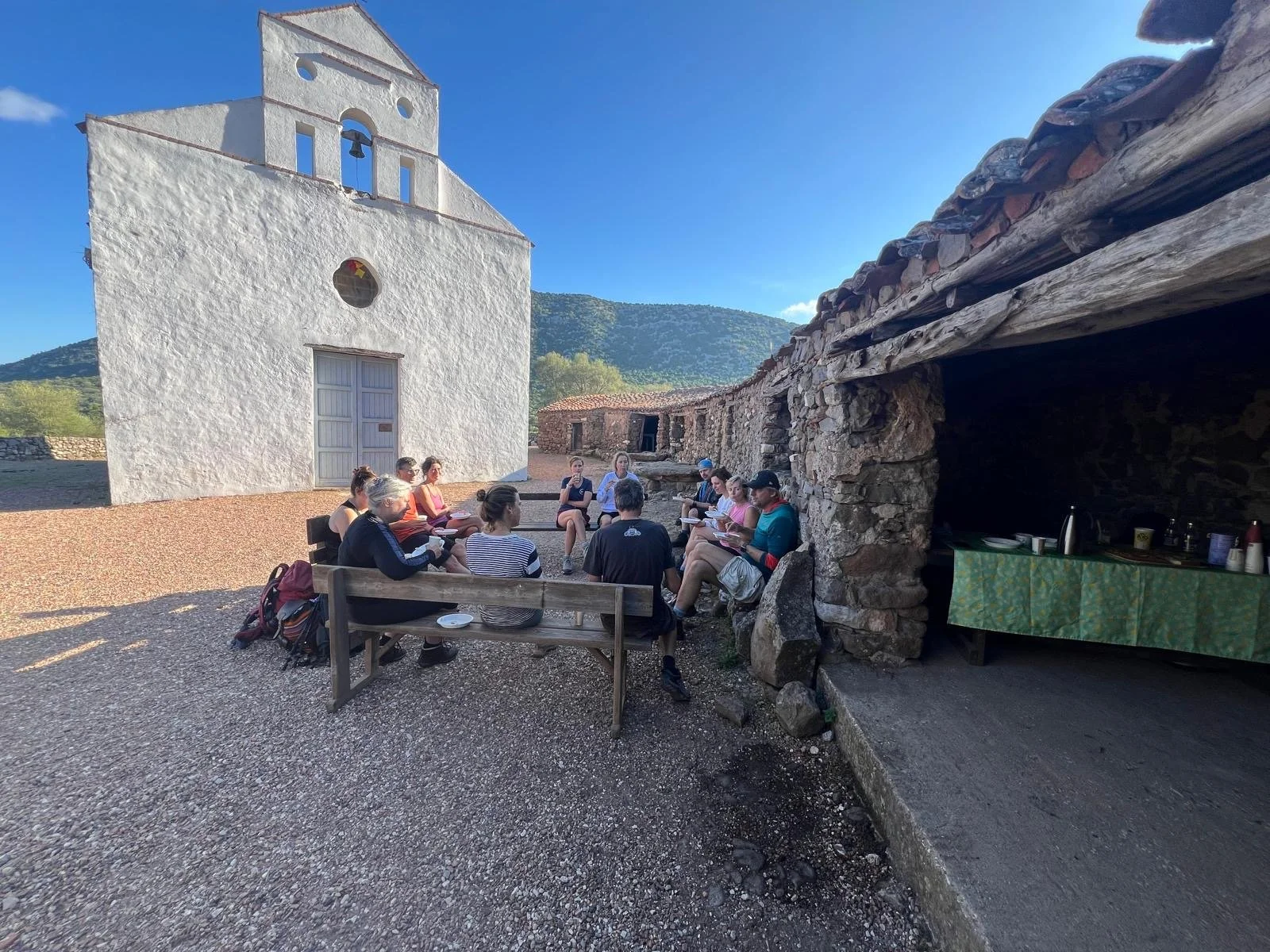 A group of people sitting in a semi-circle outdoors near an old white church and a stone shelter. The scene is set against a backdrop of hills under a clear blue sky.
