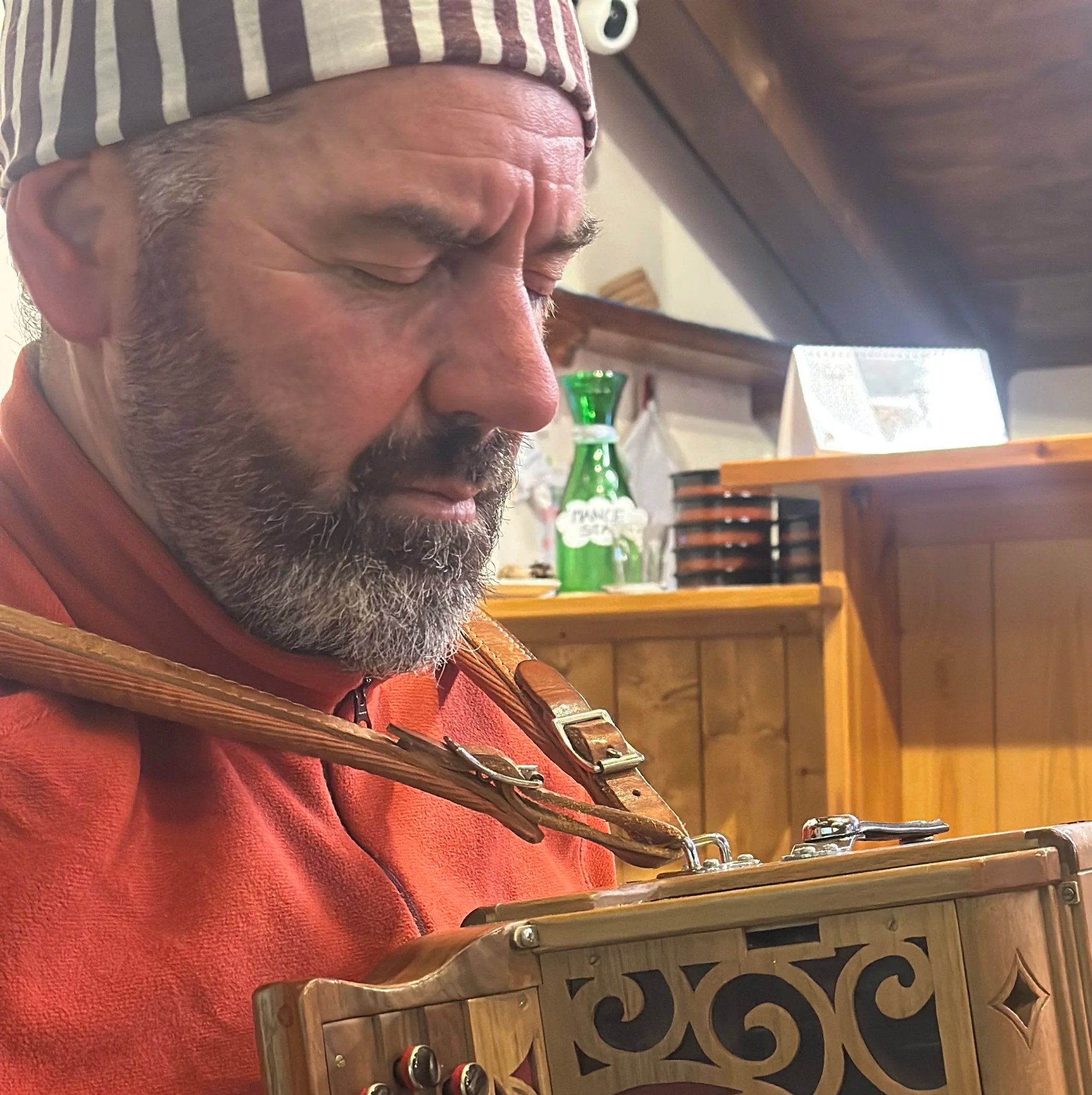 A man with a gray beard, wearing a striped hat and an orange shirt, playing a wooden accordion indoors, with wooden furniture and a green glass bottle in the background.